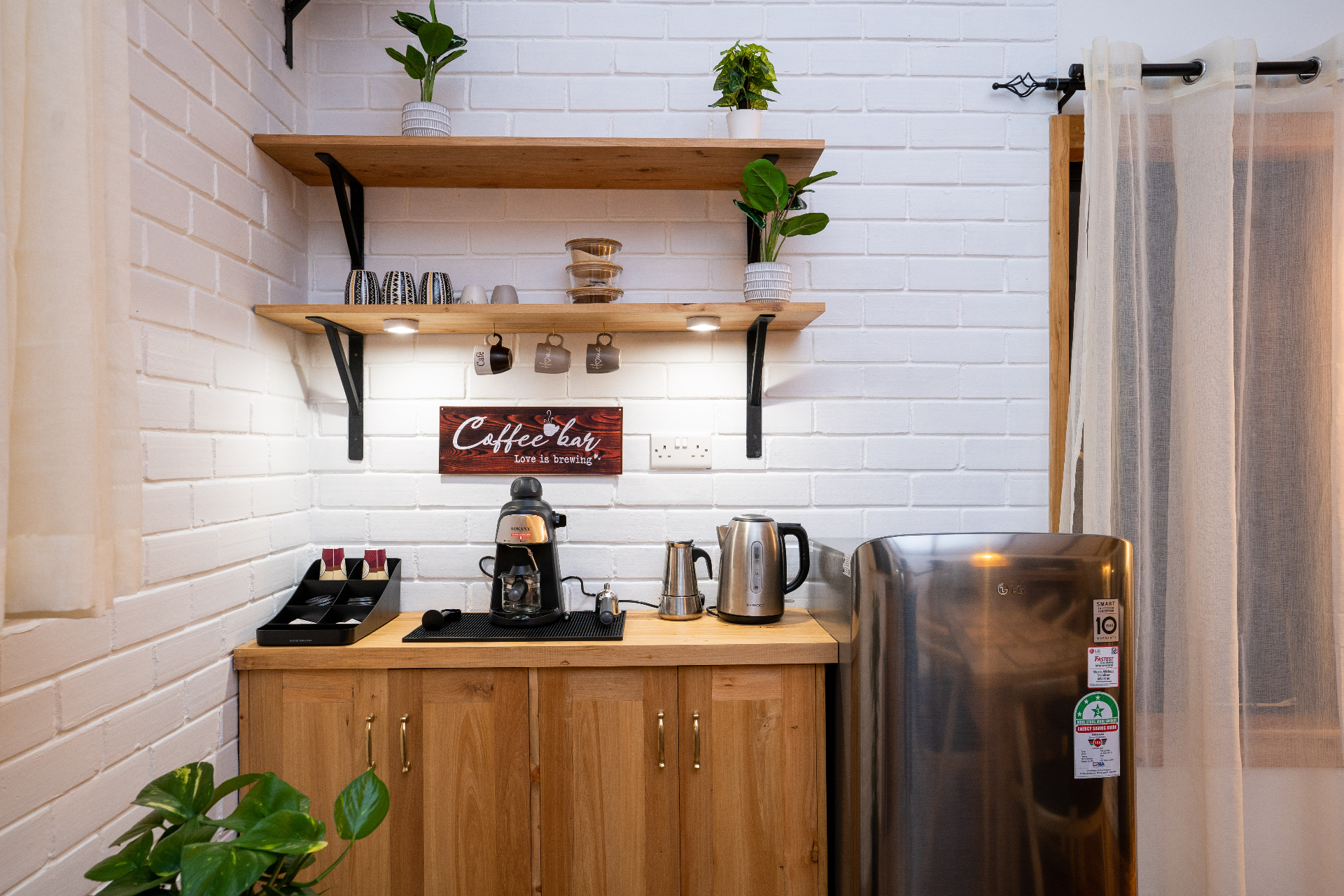 Dedicated coffee bar nook in Cottage 1 with espresso machine, kettle, mini fridge, open timber shelving, mugs and small plants against a white brick wall