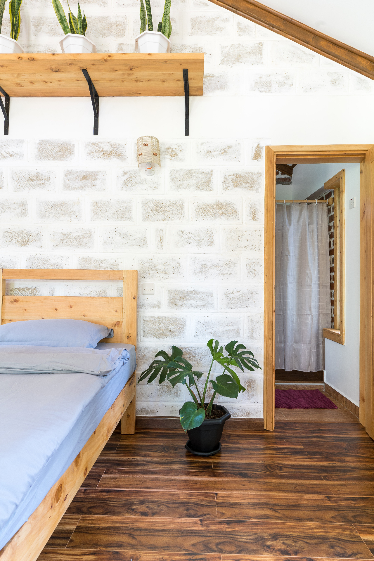 Side view from the bed toward the en suite doorway, with a monstera plant, whitewashed stone wall and timber shelf above