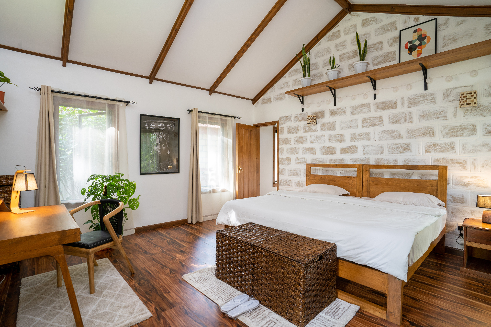 Wide view of Cottage 2 bedroom with twin beds pushed together under a vaulted white ceiling with timber beams, exposed brick feature wall, potted plants and framed African portrait