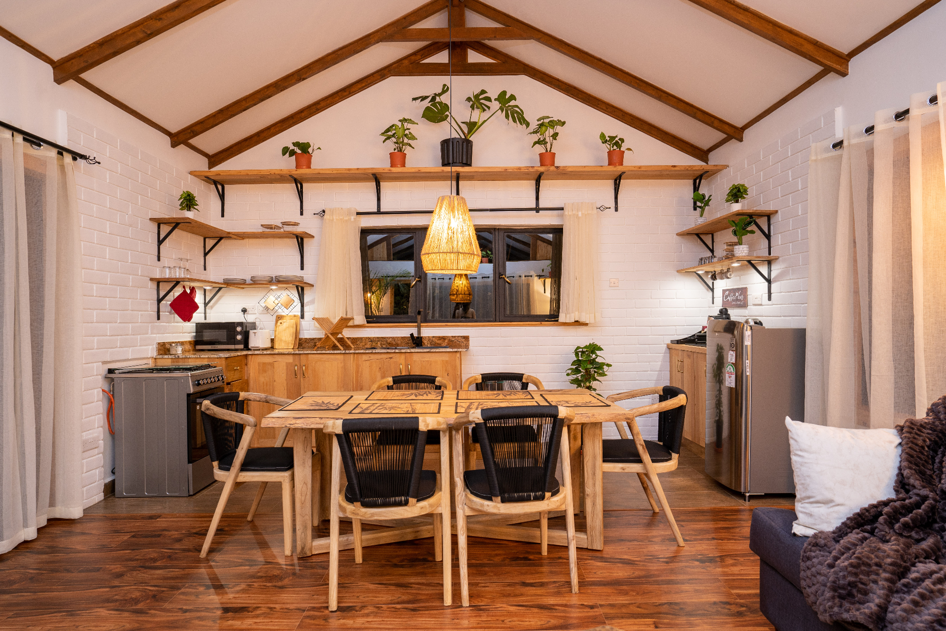 Head-on view of the wooden dining table for six framed by open shelving, potted plants and a woven pendant light above the kitchen with gas cooker and fridge