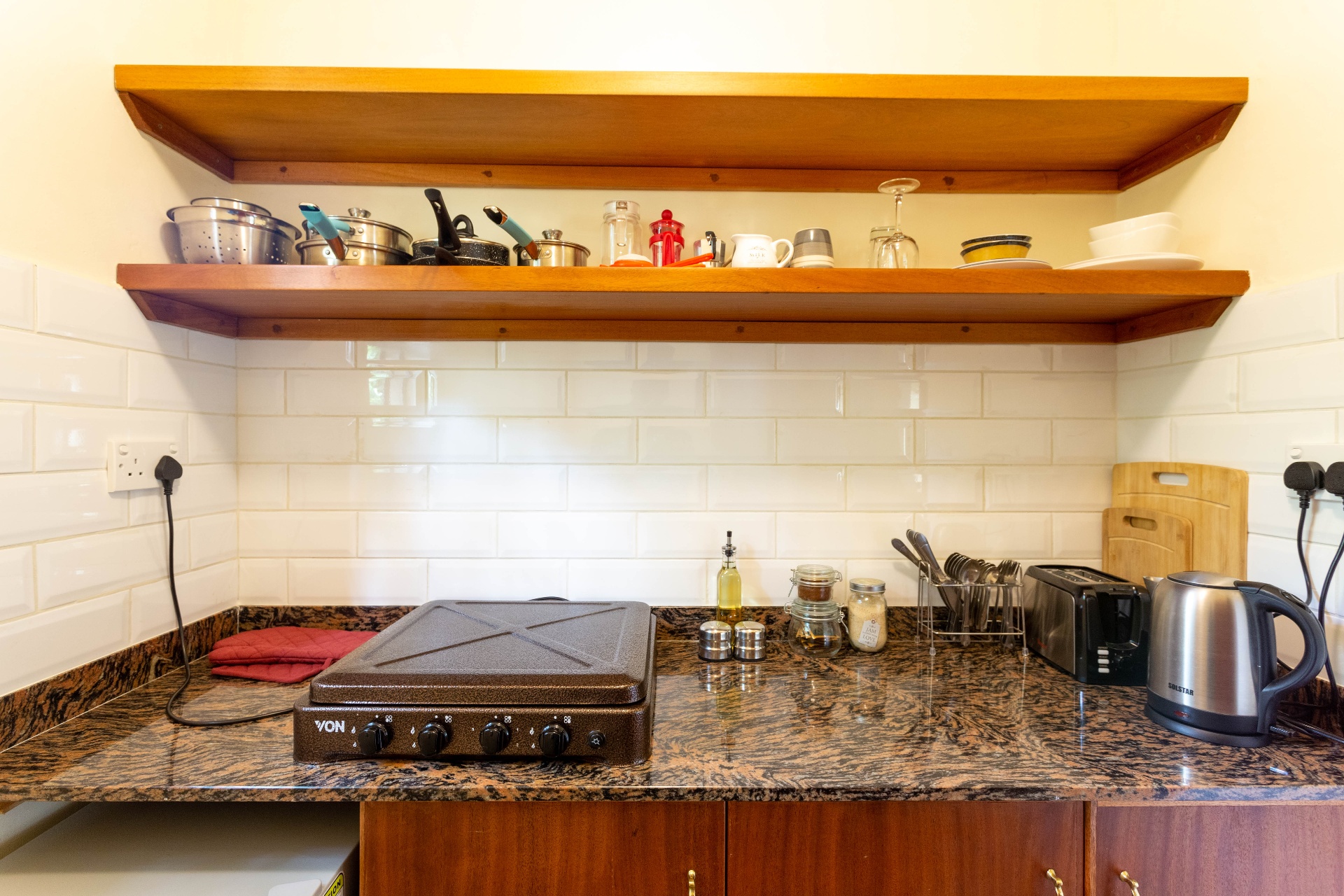 Wide view of the kitchenette with cooktop, kettle, toaster and open timber shelves stocked with cookware and glassware above a subway-tile splashback