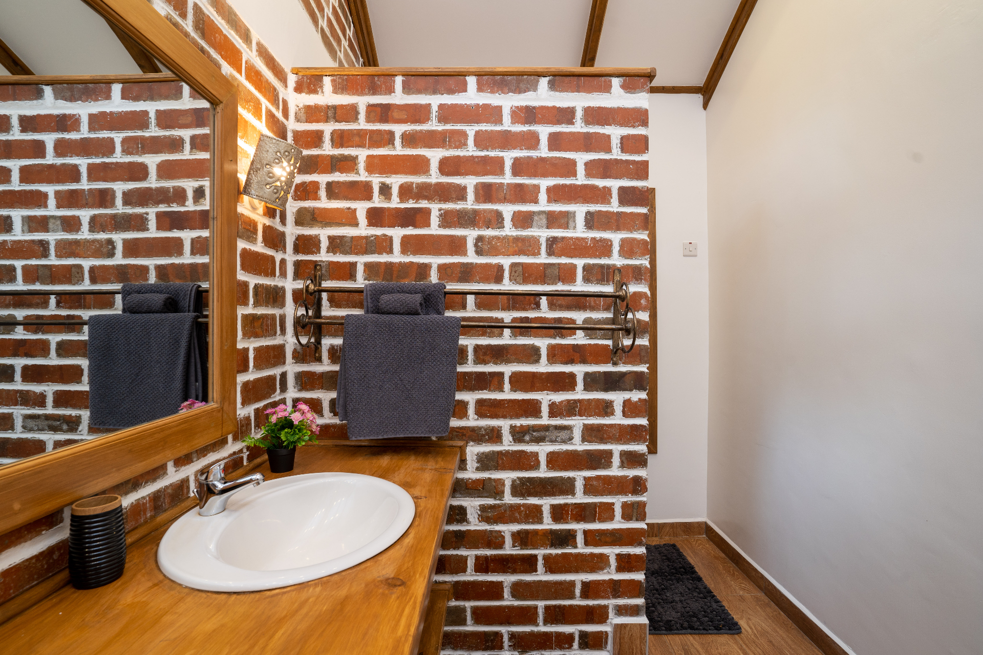 Cottage 2 bathroom with timber vanity, oval basin, framed mirror reflecting brickwork and a wrought iron towel rail with folded grey towels