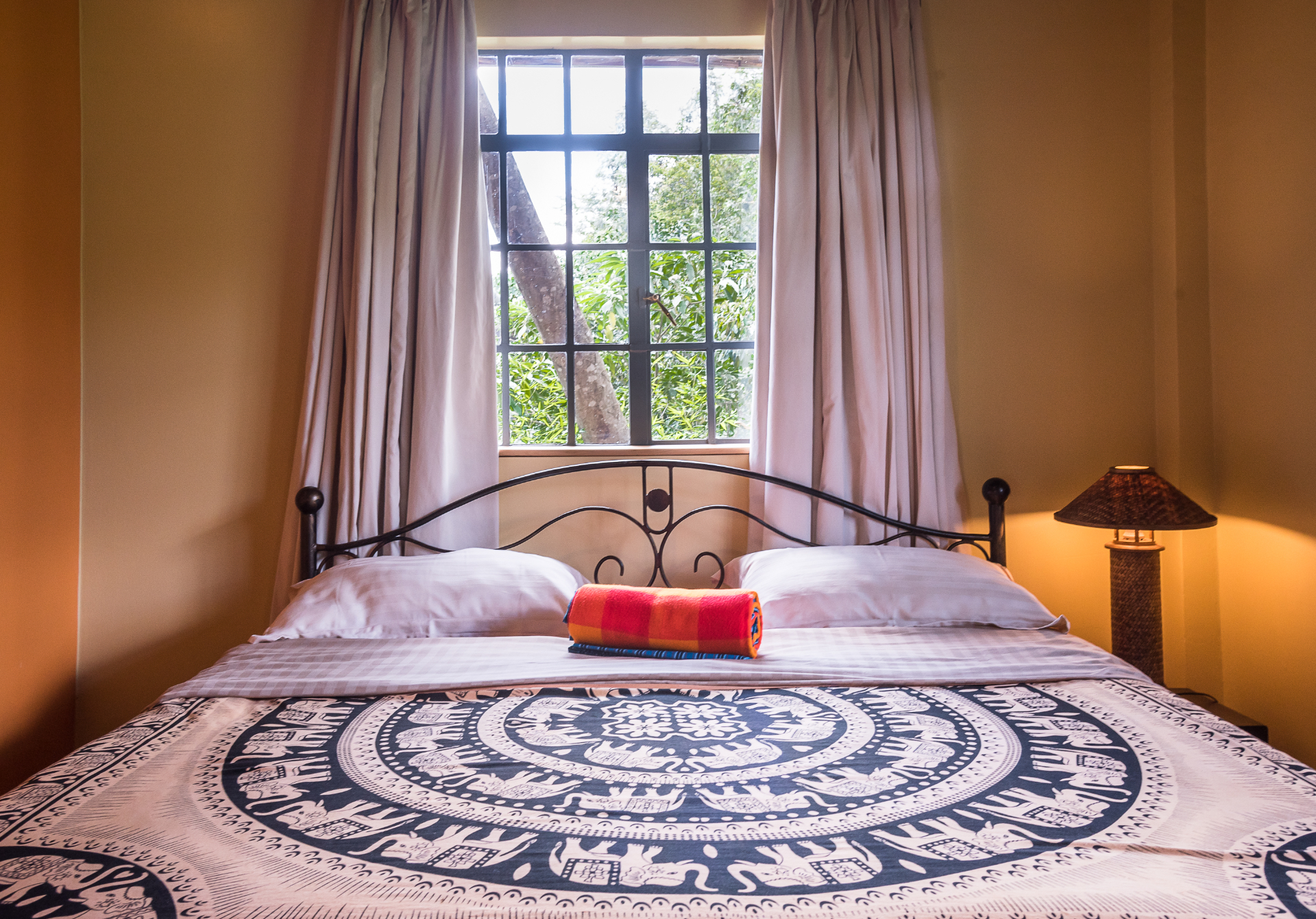 Queen bed with iron headboard and elephant-motif throw beneath a tall steel-framed window opening onto jungle foliage