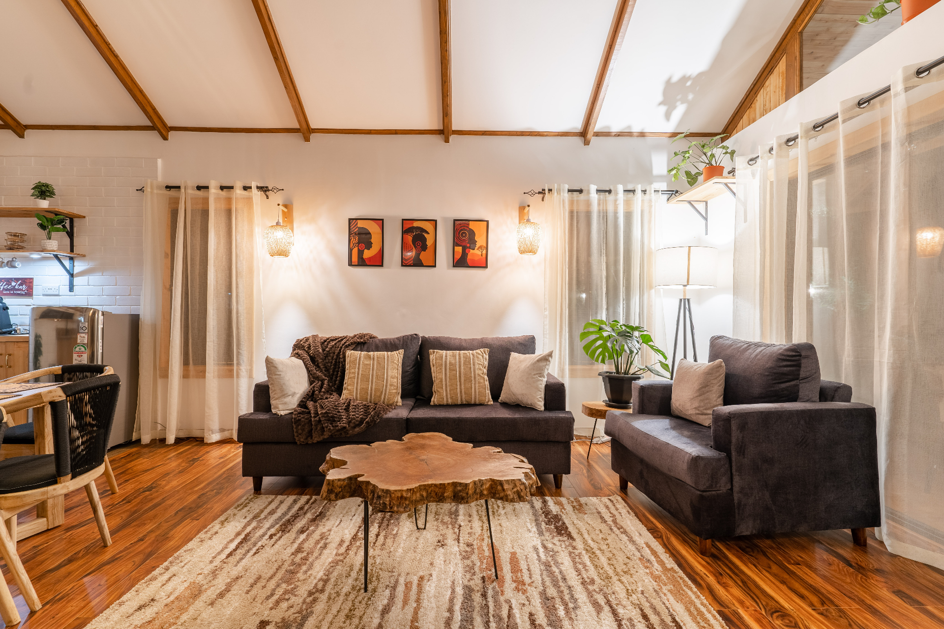 Wide view from the lounge showing the sofa and armchair, live-edge coffee table and rug in front, with the open kitchen and dining area visible to the left under a pitched wood ceiling