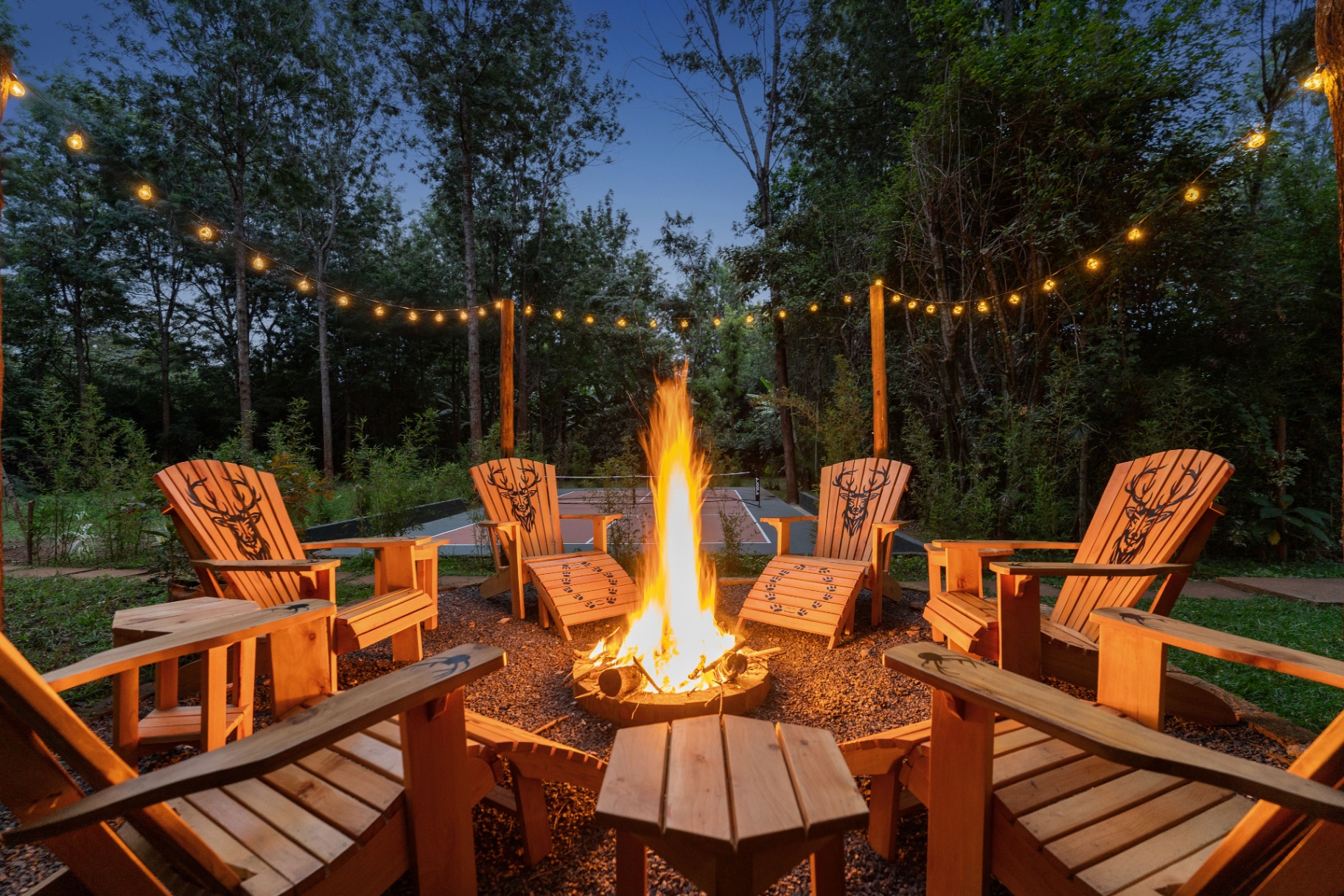 Tall flames rising from a stone firepit ringed by Adirondack chairs at dusk with the pickleball court and forest visible beyond under strung fairy lights