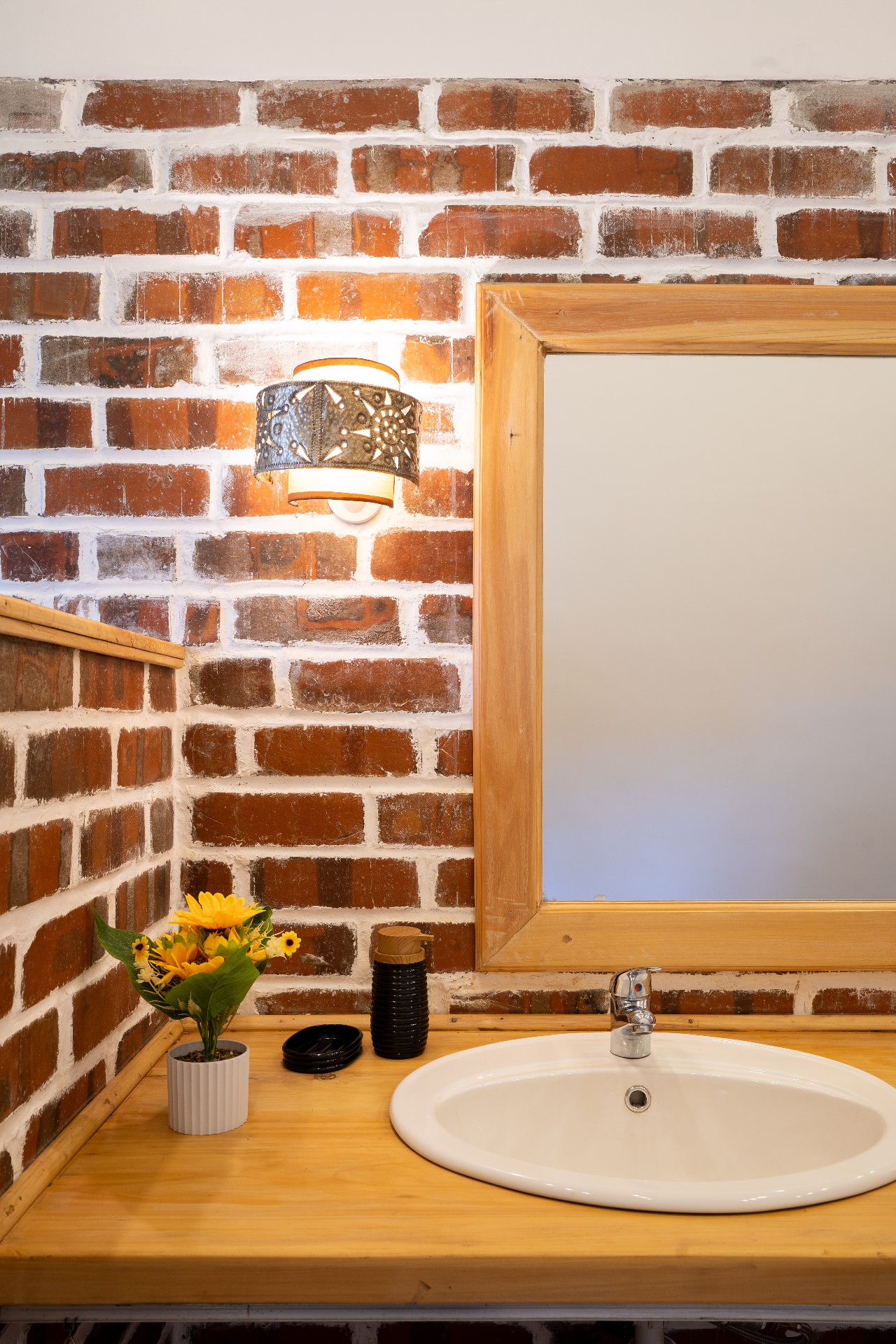Close-up of one basin, a framed mirror and a patterned metal sconce lighting the whitewashed brick corner