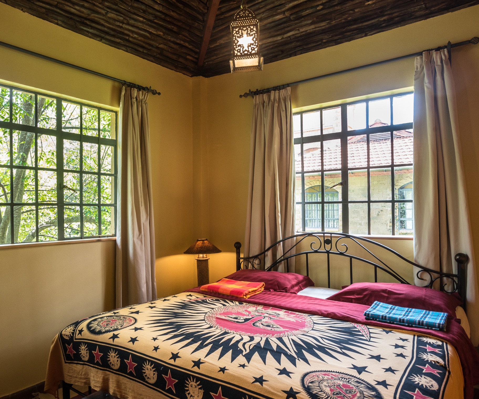 Corner bedroom with sun-and-stars tapestry bedding, Moroccan lantern and two large windows opening onto trees and terracotta rooftops