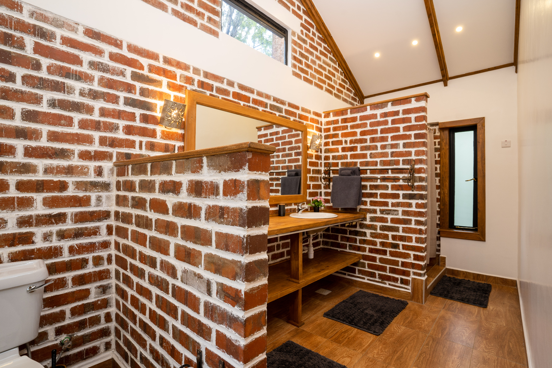 Cottage 2 bathroom with exposed red-brick partitions, timber vanity shelf, framed mirror, dark towels and warm wall sconces beneath a vaulted ceiling
