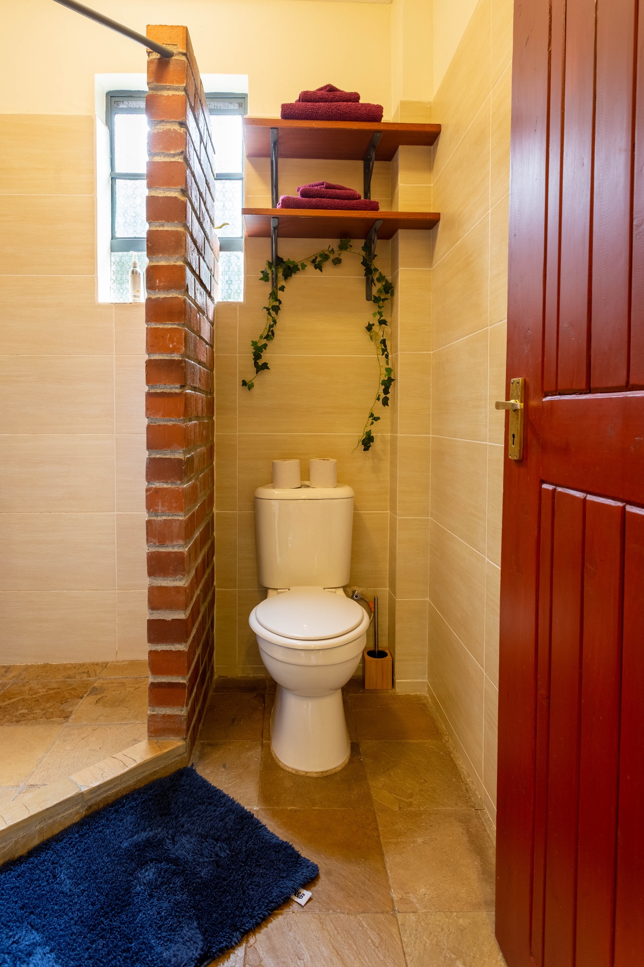 Bathroom WC nook with exposed-brick partition, folded burgundy towels on timber shelves and trailing ivy