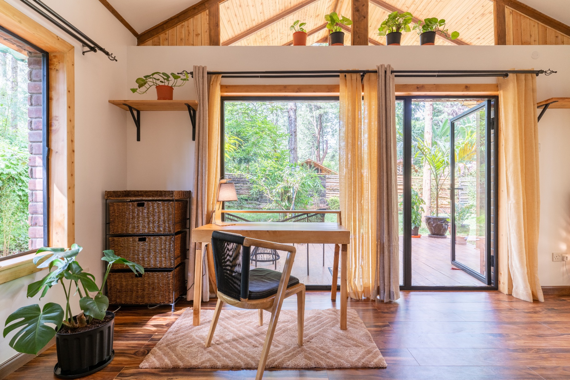 Writing desk and chair in front of a wide window and open terrace door leading onto a private deck surrounded by tropical greenery