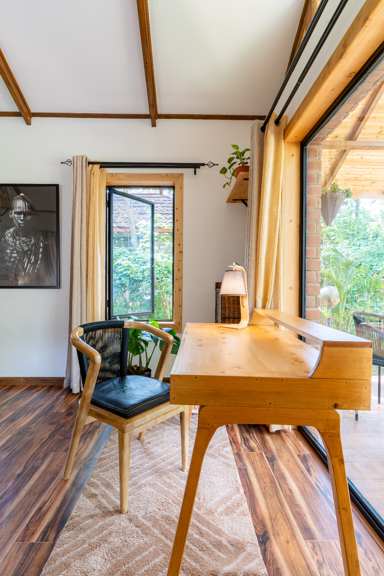Timber writing desk and rope-backed chair beside an open casement window, framed portrait on the wall and terrace doors letting in garden light