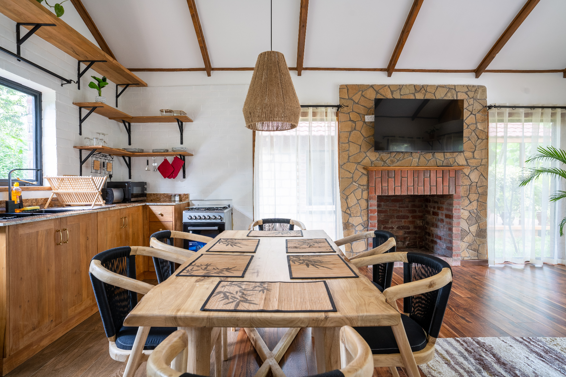 Cottage 2 dining area with a timber table for six beside the kitchen, facing a stone-clad fireplace with mounted television under the vaulted timber-beam ceiling