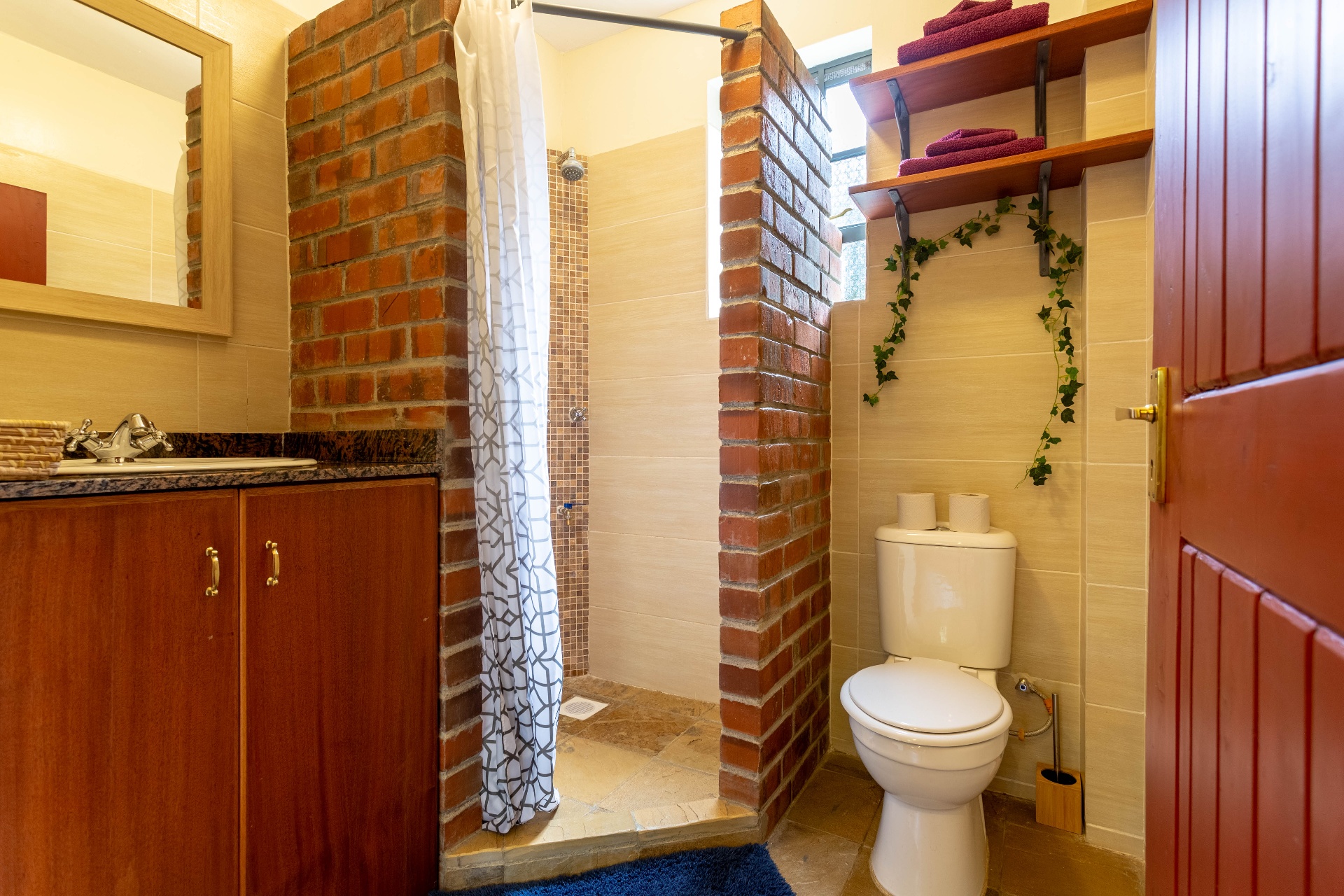 En suite bathroom with exposed-brick walk-in shower divider, timber vanity, WC and shelved towels beside a red timber door