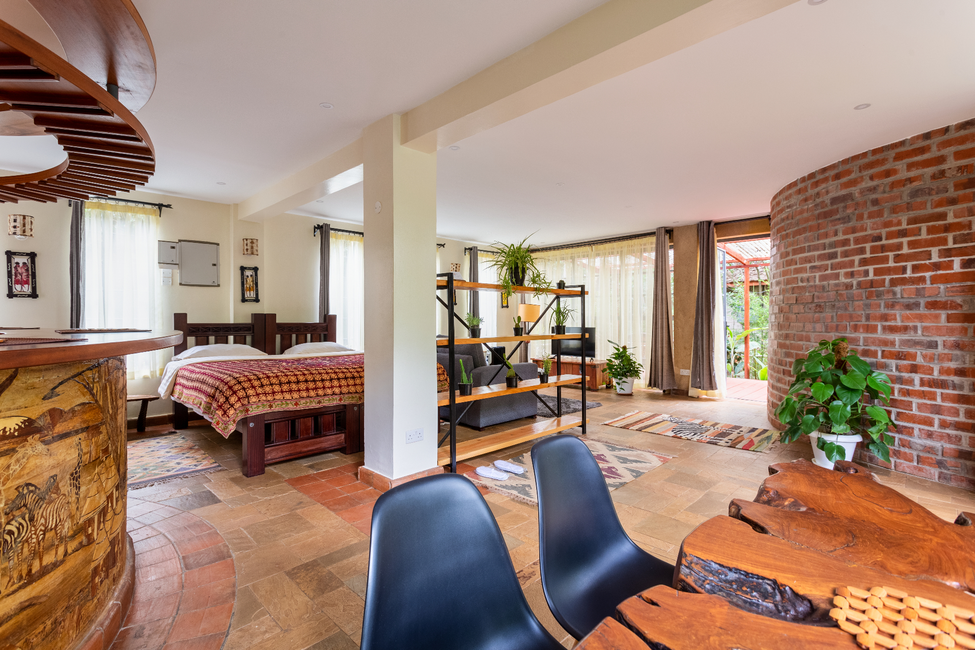 Wide view of the studio showing the carved timber bed, lounge with plant-shelf divider, curved brick wall and terrace doors