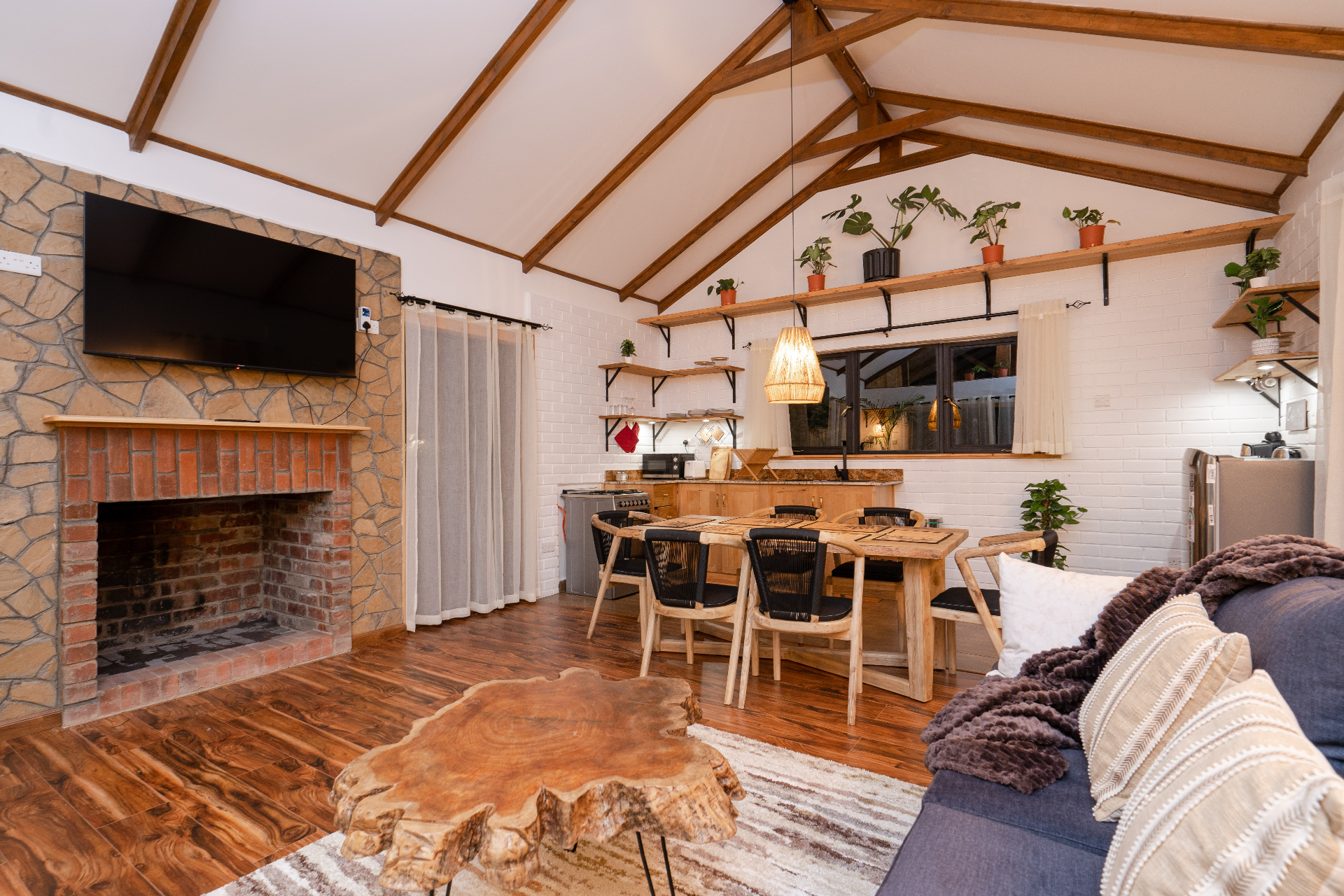 Wide interior view of Cottage 1 featuring the stone fireplace with mounted TV, live-edge coffee table, sofa, dining table and kitchen under the high timber ceiling