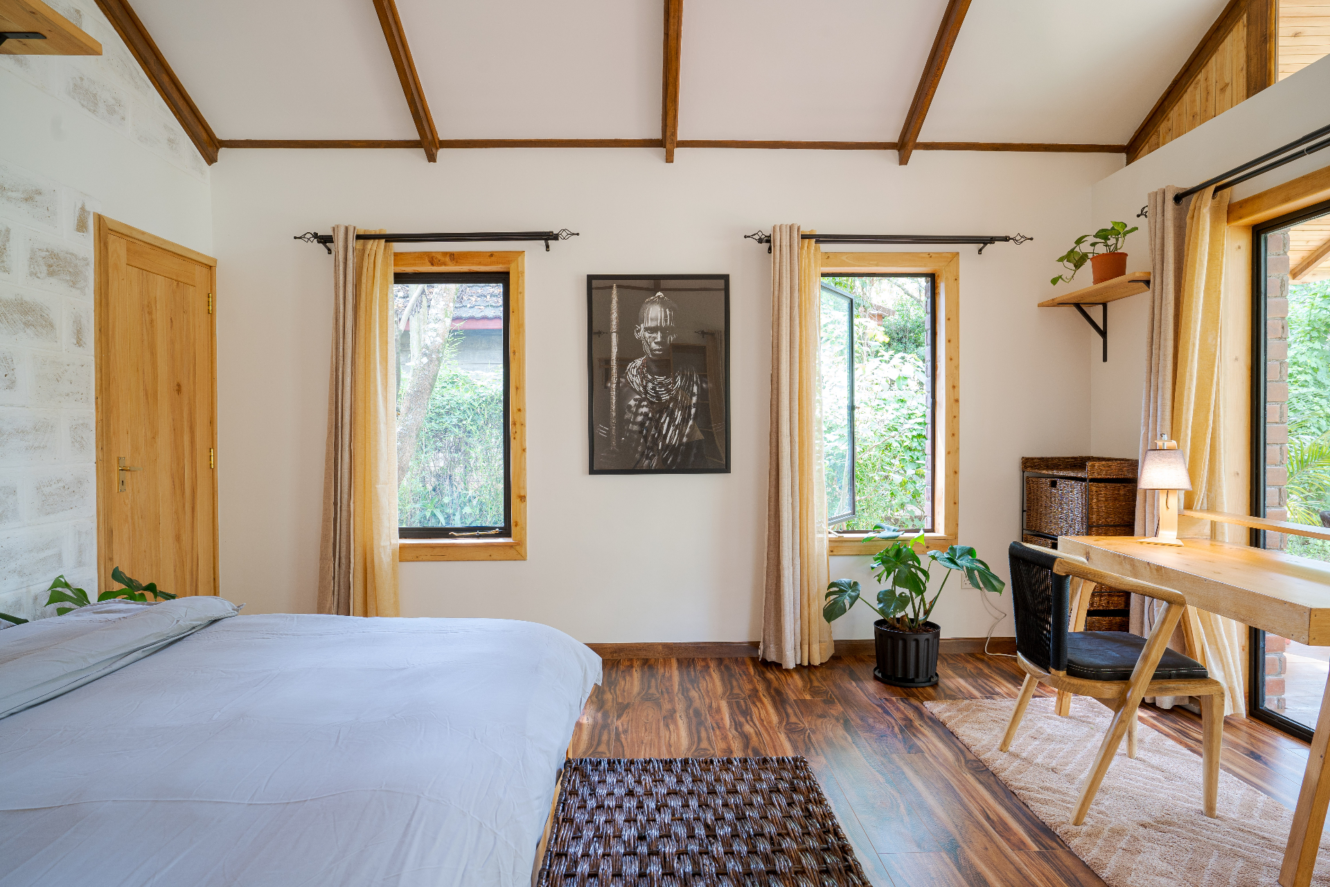 Wide bedroom view with king bed, framed African portrait between two garden-facing windows, and the writing desk under the vaulted timber ceiling