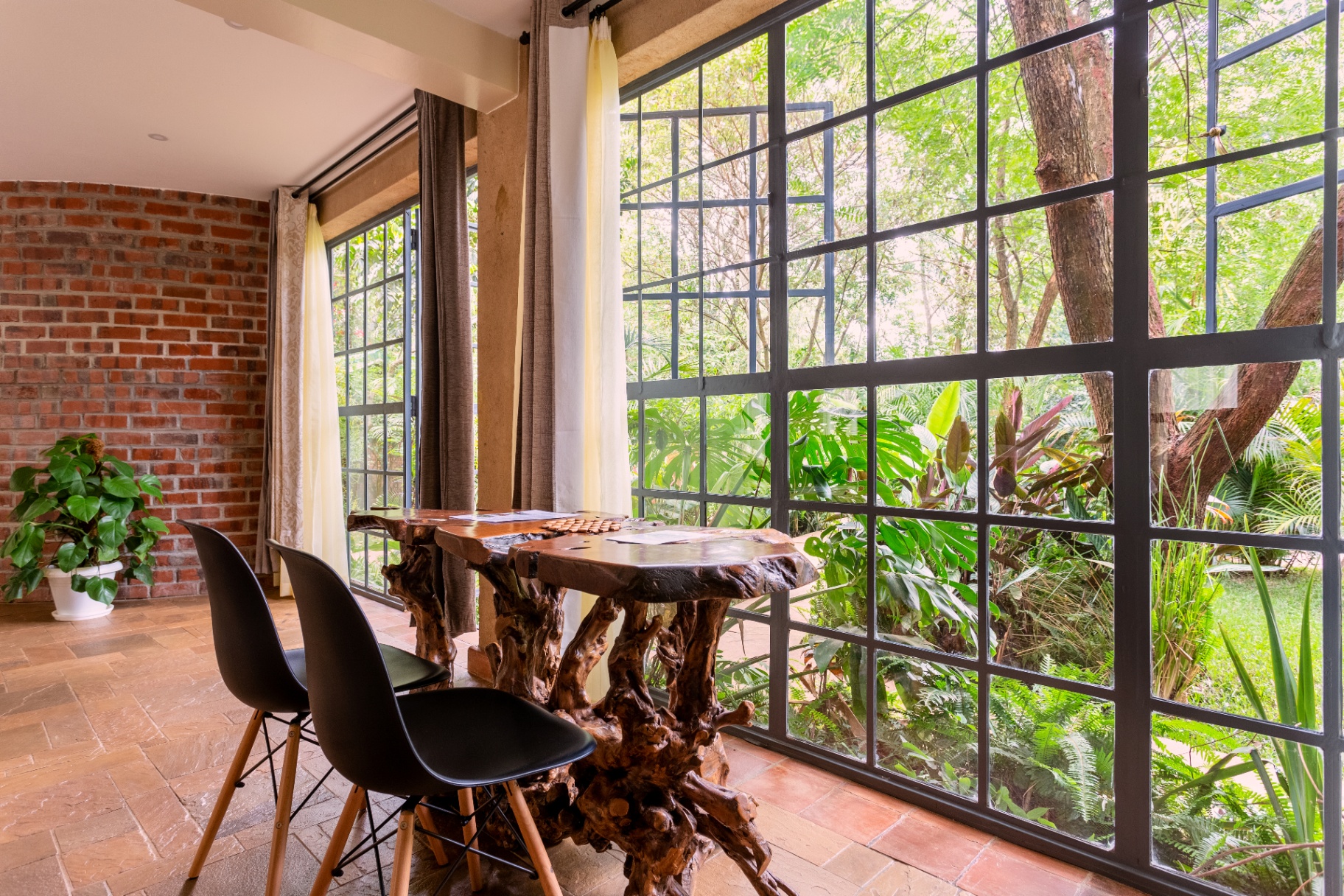 Root-carved dining table and black chairs set against a full-height steel-framed window wall overlooking the jungle garden, with a red-brick feature wall opposite