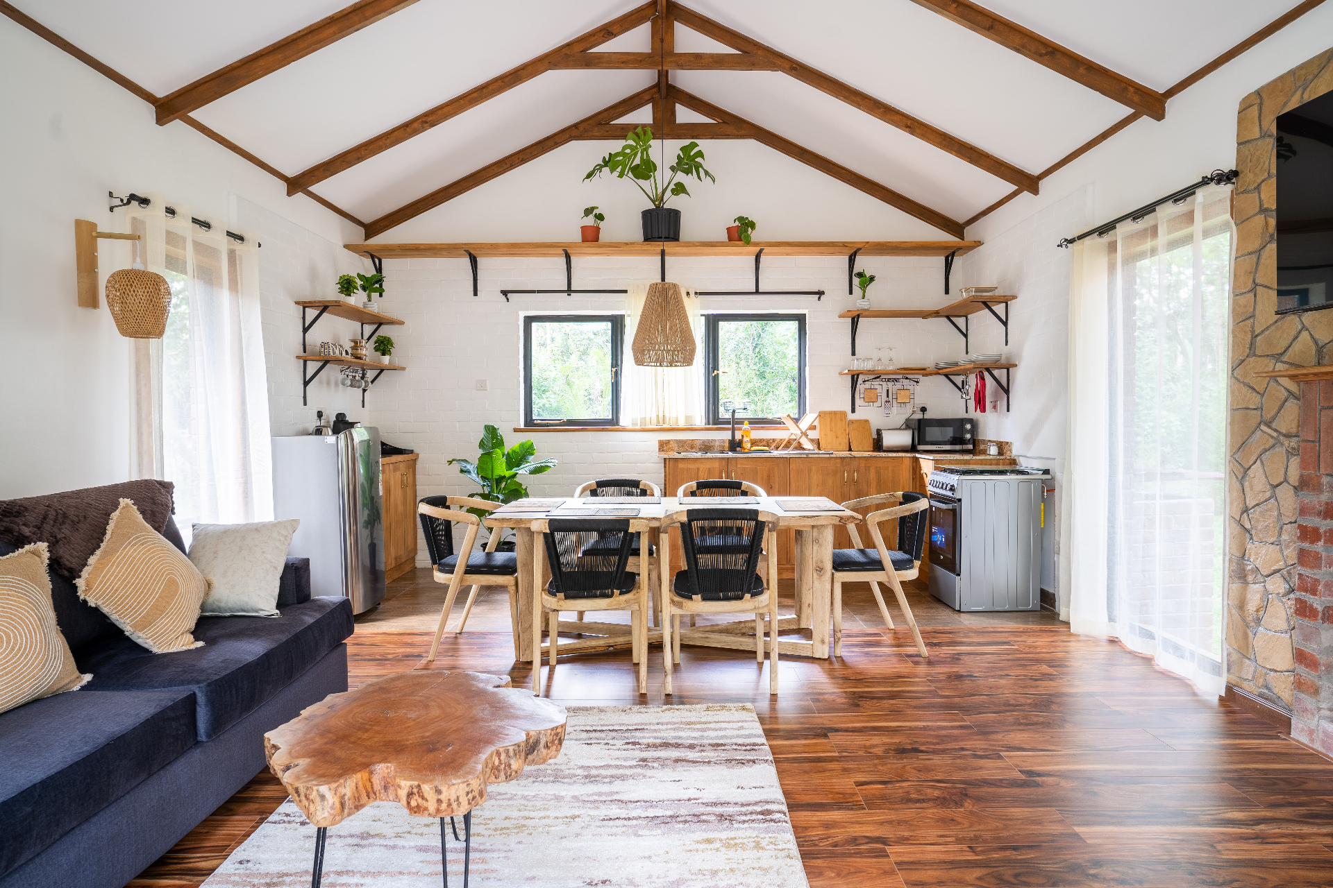Wide view of Cottage 2 open-plan living area with vaulted timber-beam ceiling, navy sofa, live-edge coffee table, dining table for six and a kitchen with open shelves and gas range