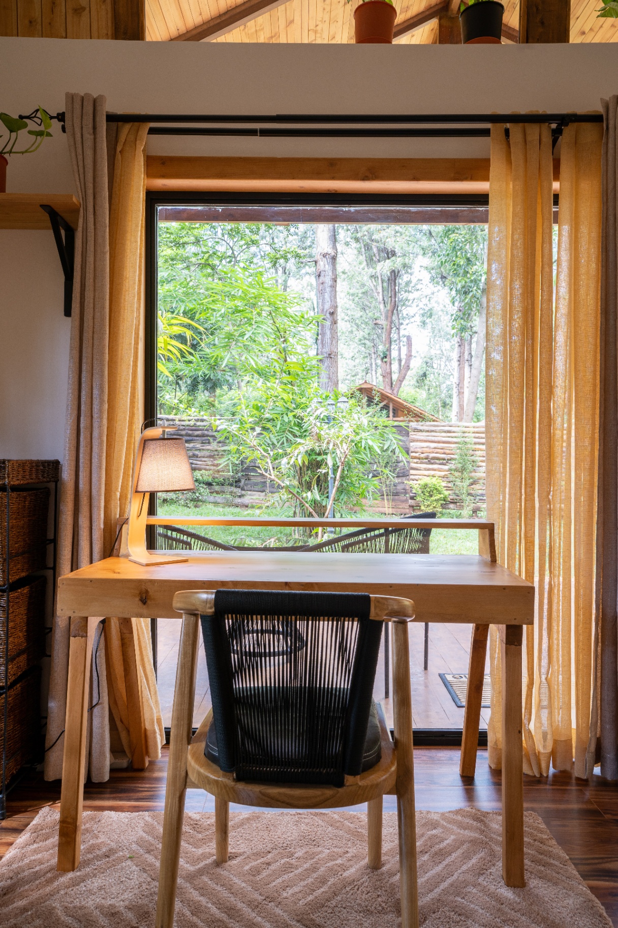 Head-on view of the timber desk and lamp framed by long curtains, looking out through a large window to bamboo and forest cottages beyond