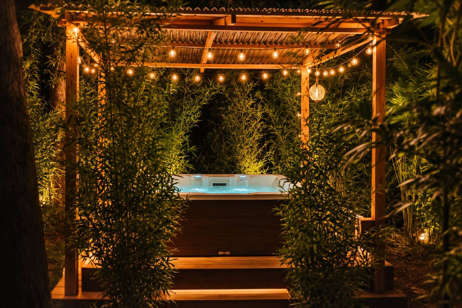 Private jacuzzi glowing turquoise at night beneath a timber pergola strung with warm festoon lights, surrounded by dense jungle greenery at Cottage 2