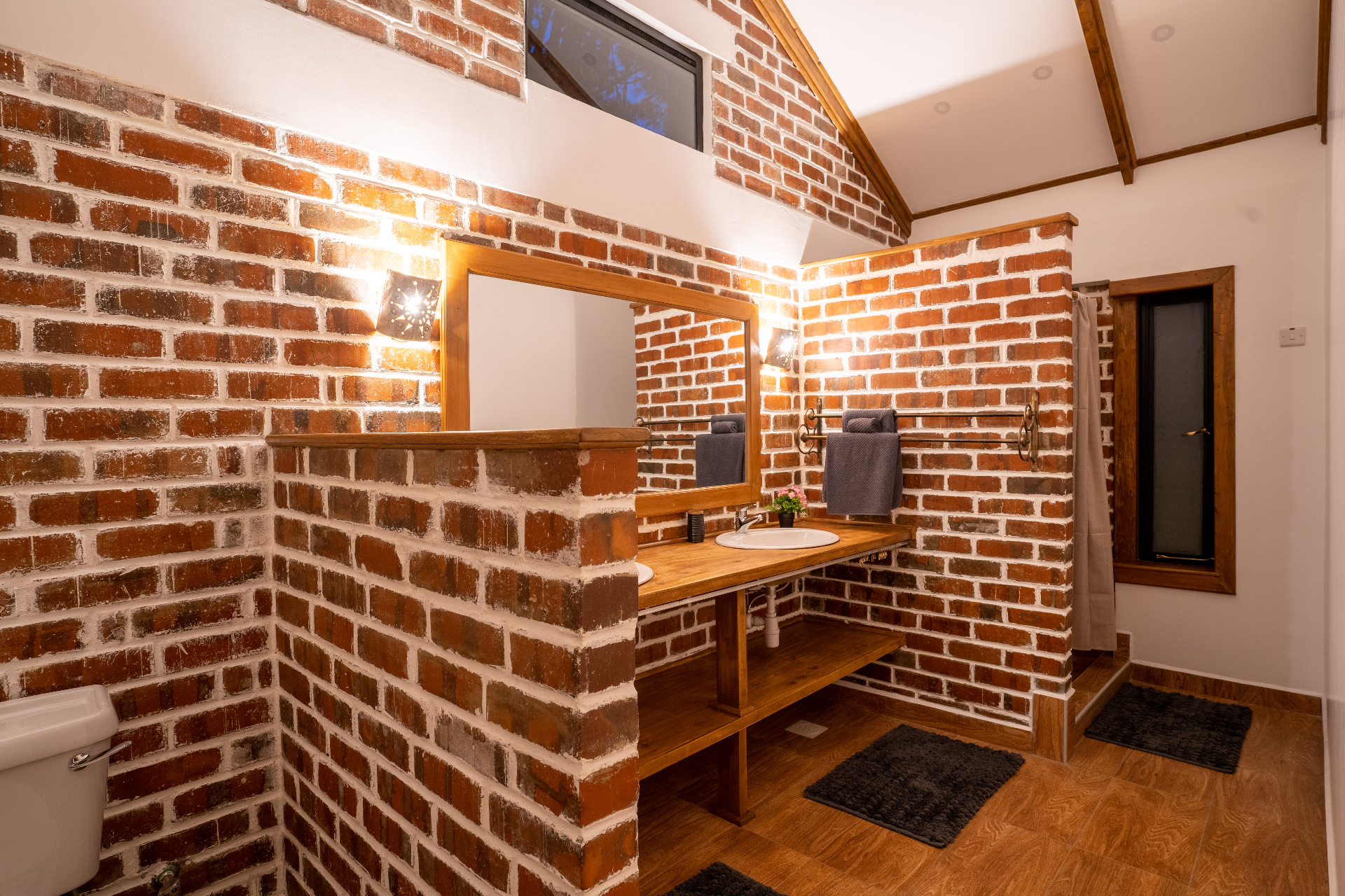 Wide nighttime view of Cottage 2 bathroom with exposed red-brick walls, double timber vanity, glowing sconces, dark bath mats and a frosted window beside the shower area