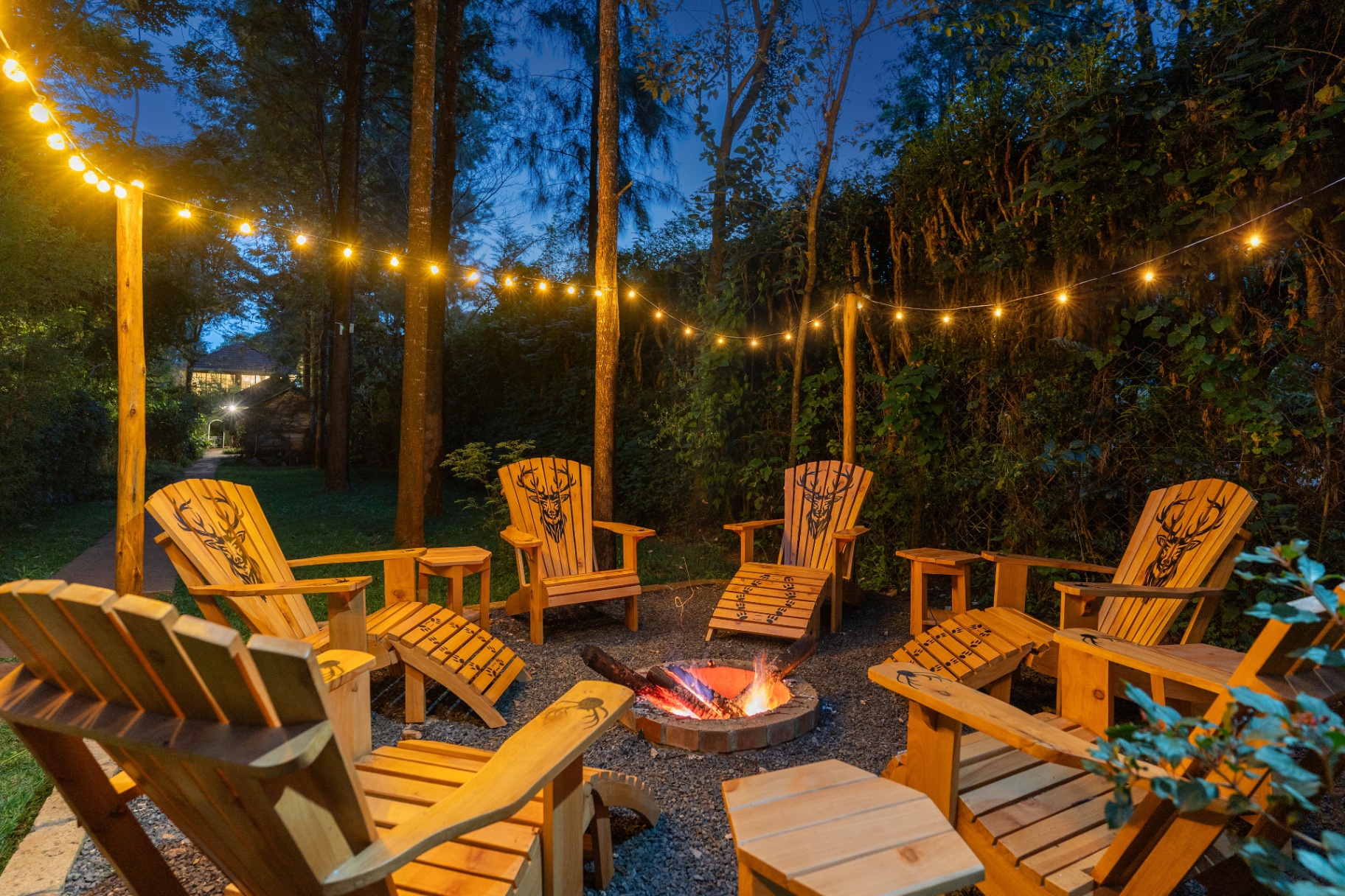 Wooden Adirondack chairs with carved stag motifs arranged around a burning firepit beneath canopies of bistro lights on a warm evening in the garden