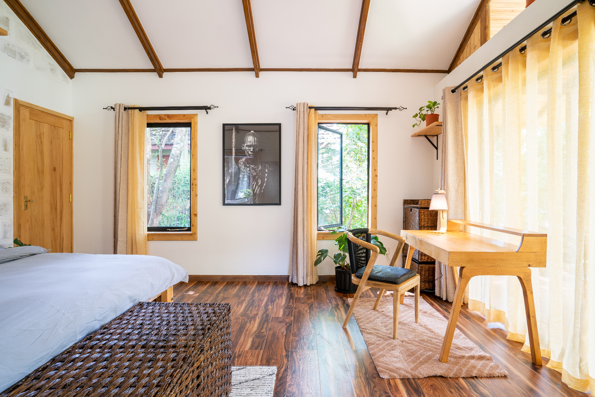 Timber writing desk and chair beside floor-to-ceiling curtains and an open window, with the bed just out of frame to the left