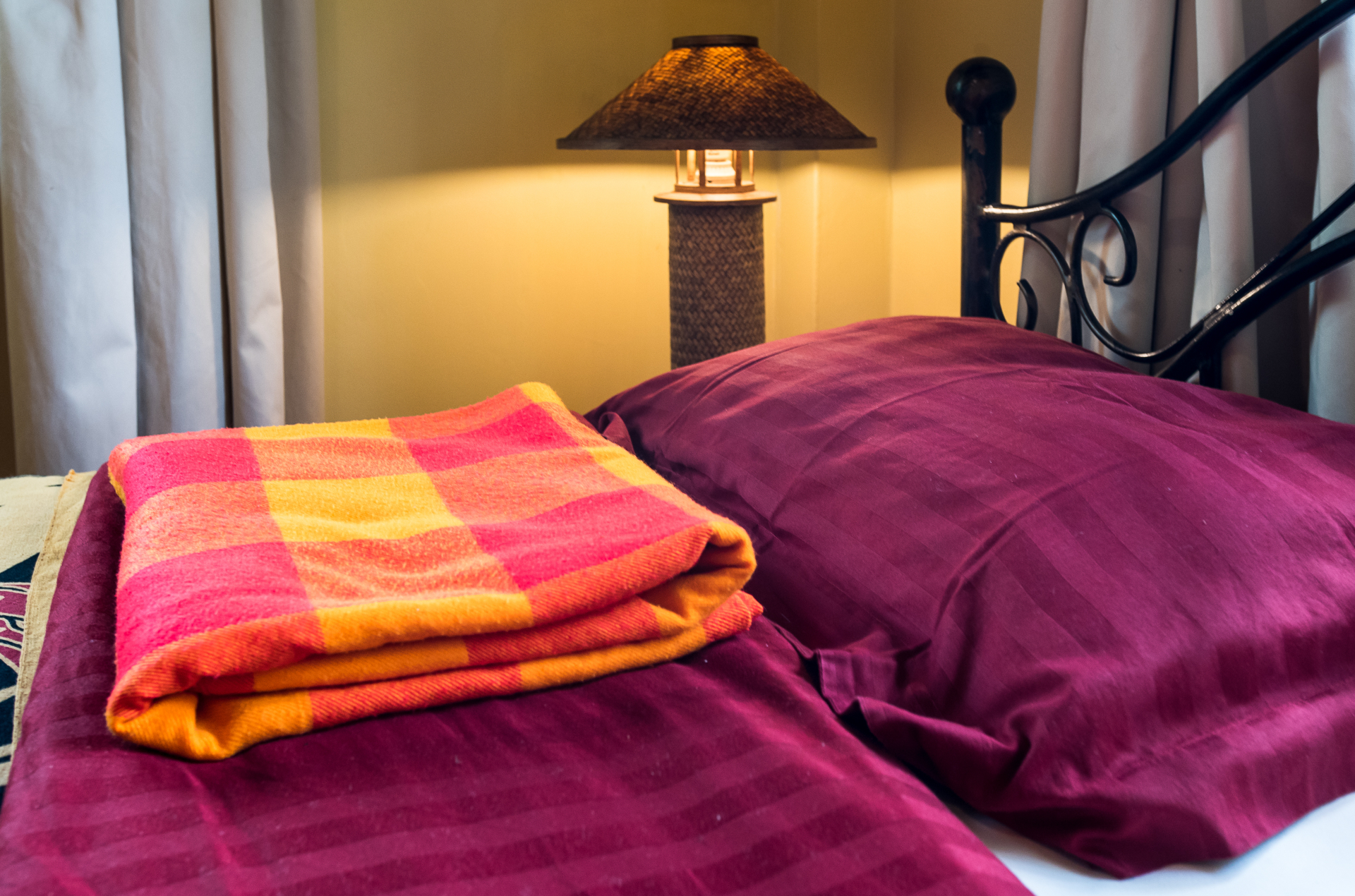 Close-up of the bed with deep plum pillows, a colourful folded blanket and a woven bedside lamp