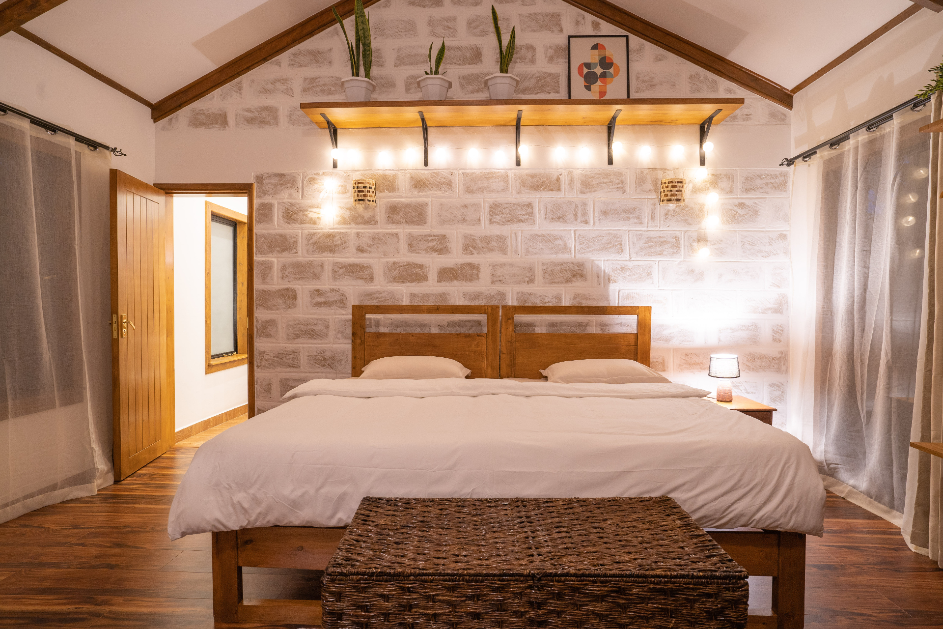 Head-on nighttime view of the Cottage 2 bed with white linens, woven trunk bench, bedside lamp and string lights glowing beneath a shelf on the stone feature wall