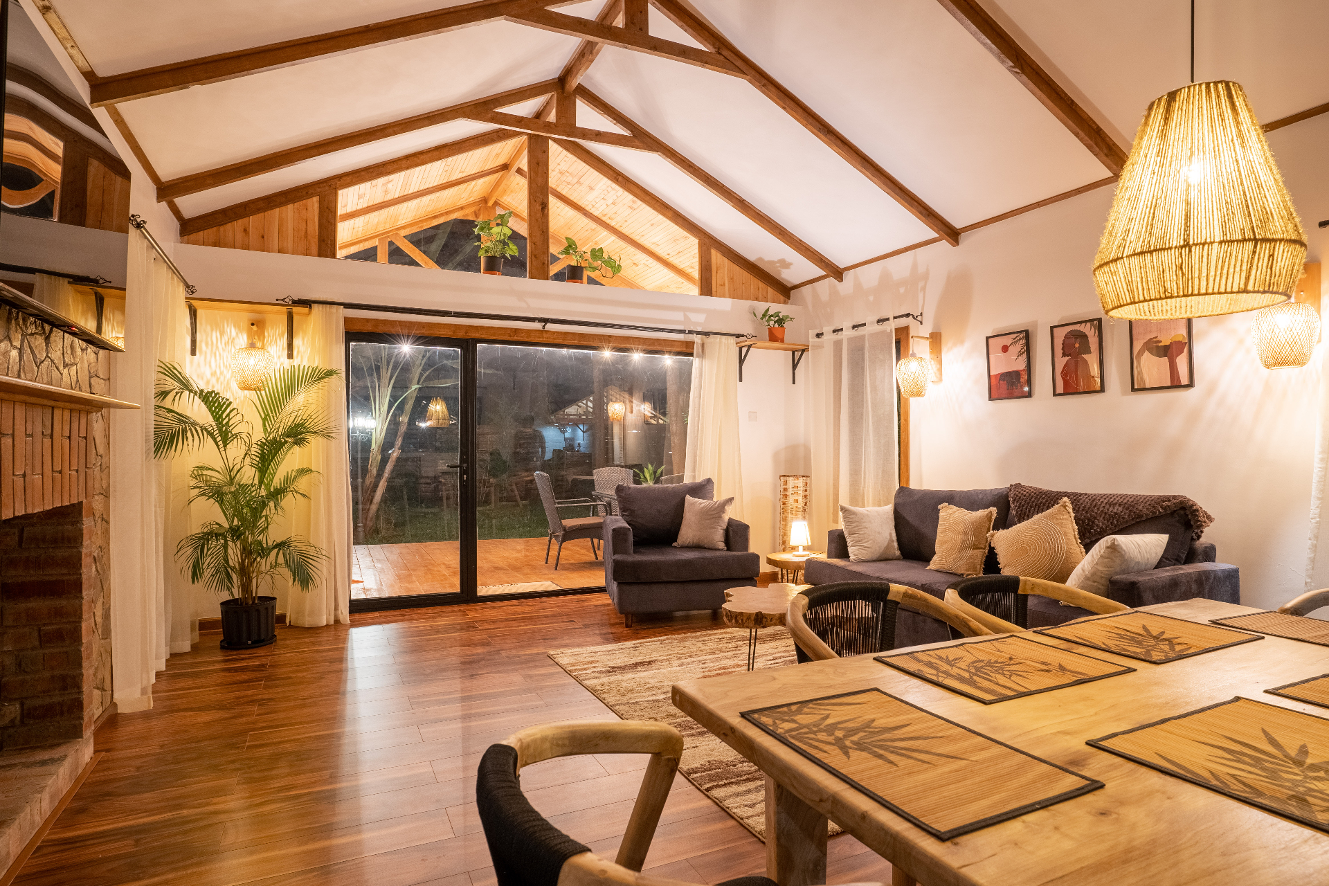 Warm evening view across Cottage 2 showing the dining table, sofa, armchair, terrace doors and vaulted timber ceiling lit by woven pendant and wall lamps