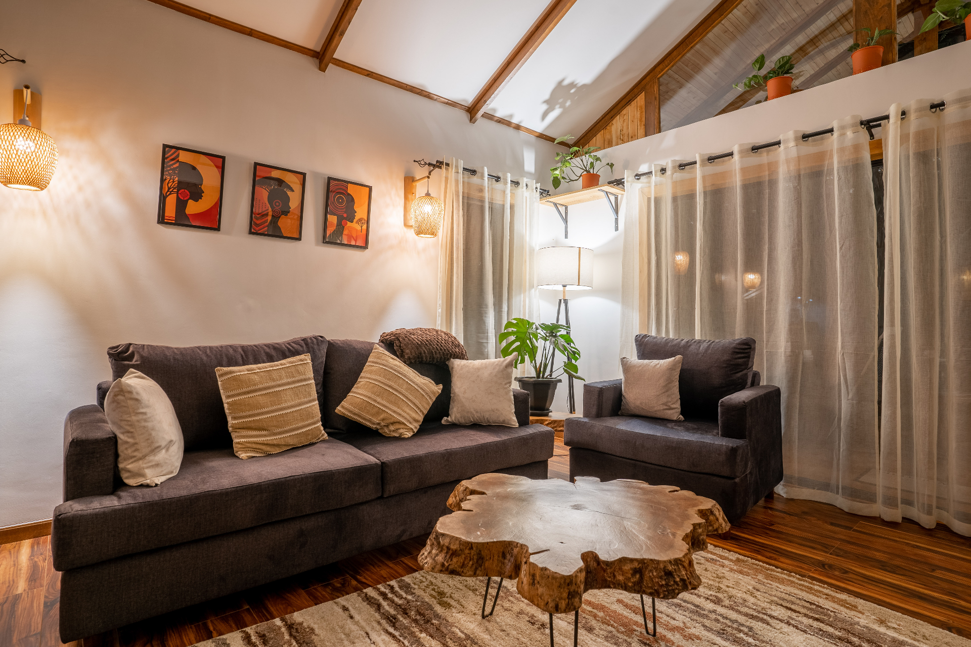 Warmly lit lounge in Cottage 1 with a charcoal sofa, matching armchair, live-edge tree slice coffee table and three orange African portrait prints on the wall under a vaulted timber-beamed ceiling
