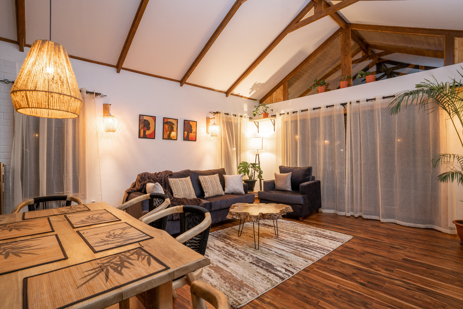 Evening view across the dining table into the Cottage 1 lounge with sofa, armchair, live-edge coffee table and softly lit woven pendant lights beneath the vaulted ceiling
