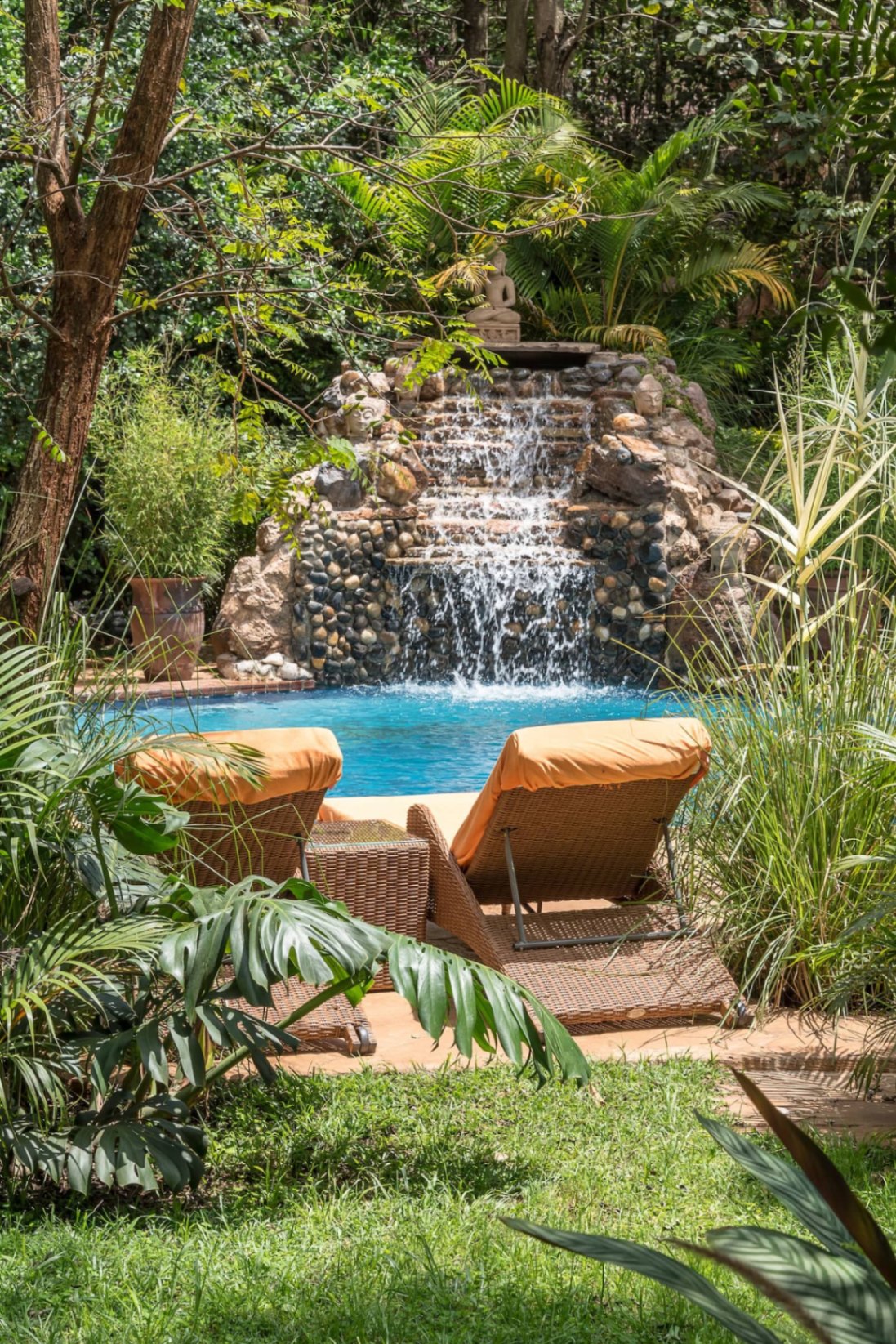 Two wicker loungers with orange cushions on a sunlit terrace facing the stone waterfall pool framed by tropical palms, monstera and lush foliage