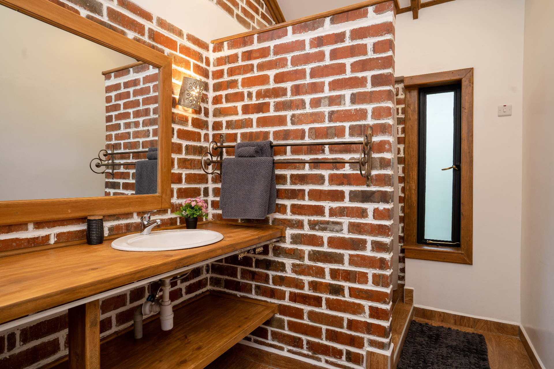 Cottage 2 en suite corner view with timber vanity, framed mirror, iron towel rail with folded grey towels against exposed red-brick walls and a slim frosted window