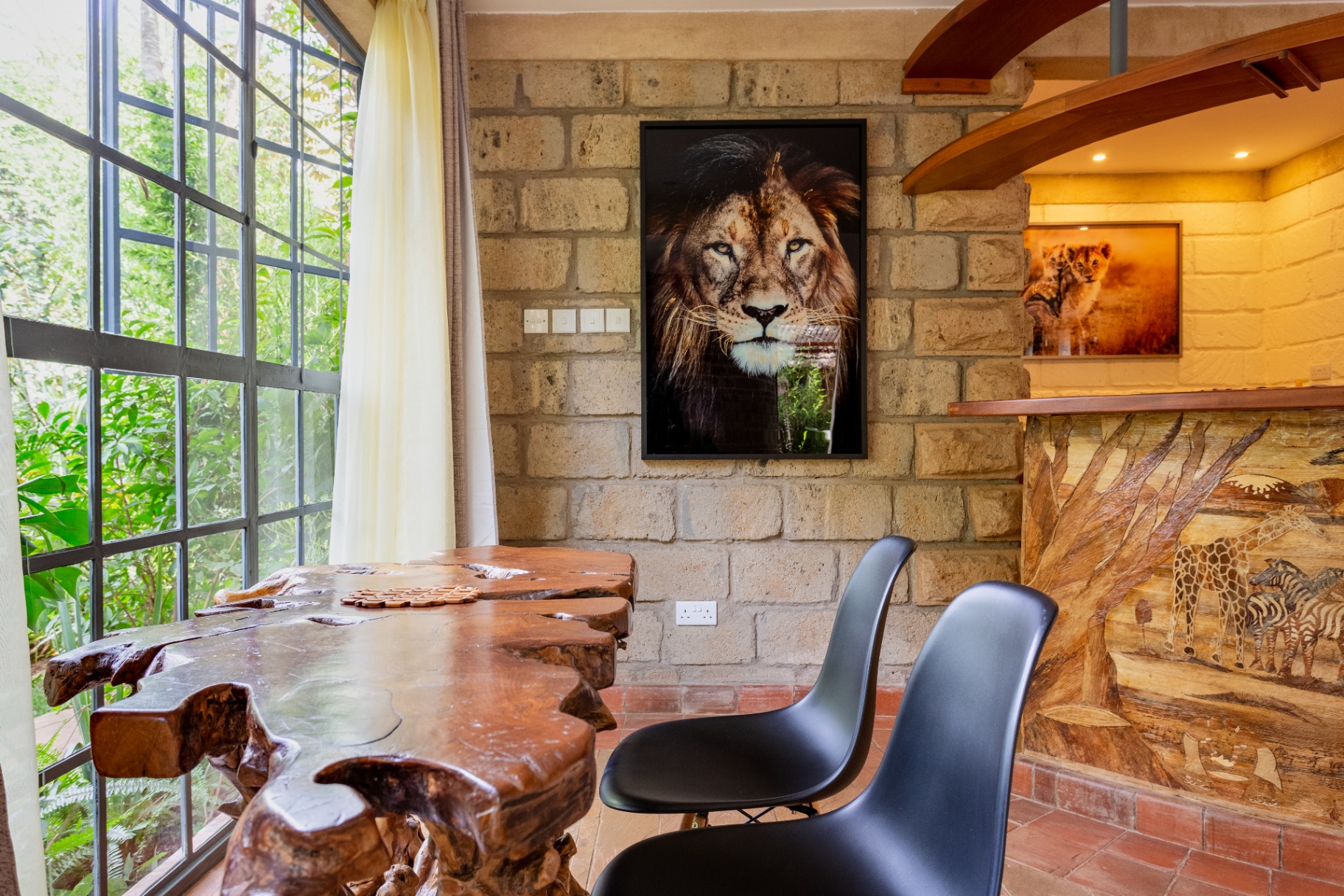 Root-carved dining table and black chairs with a framed lion portrait on the stone wall and carved safari bar beside