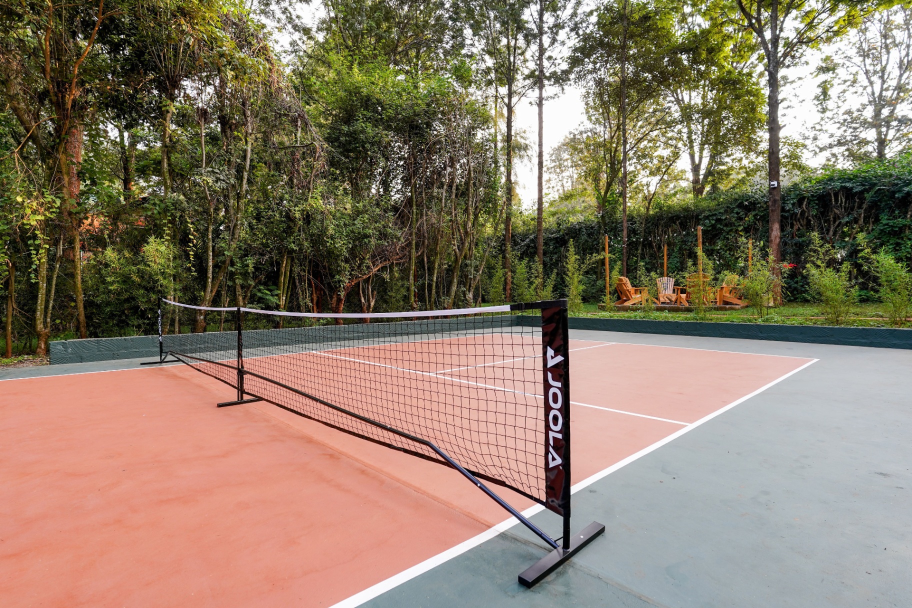 Terracotta and green pickleball court with JOOLA net set in a forest clearing, Adirondack firepit lounge visible through the trees at the far end