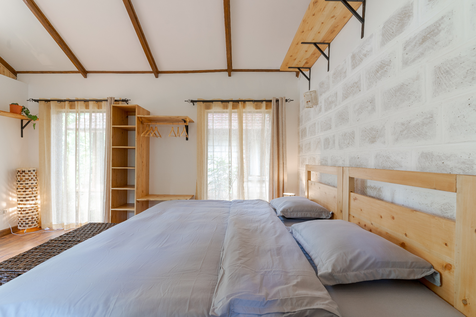 King bed with grey linens beside an open timber wardrobe with hangers, sheer curtains and a whitewashed brick accent wall under exposed rafters