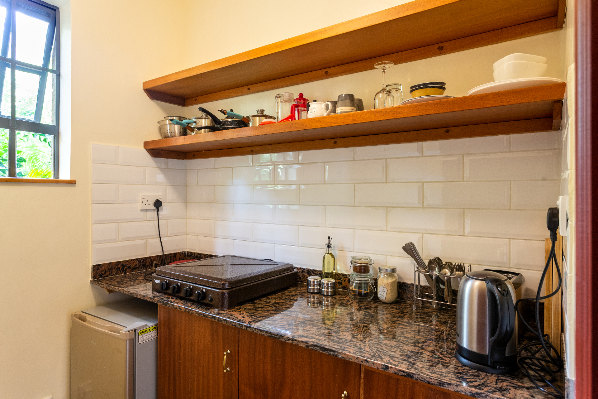 Granite worktop with electric cooktop, kettle, utensils and warm timber open shelving above a subway-tile backsplash