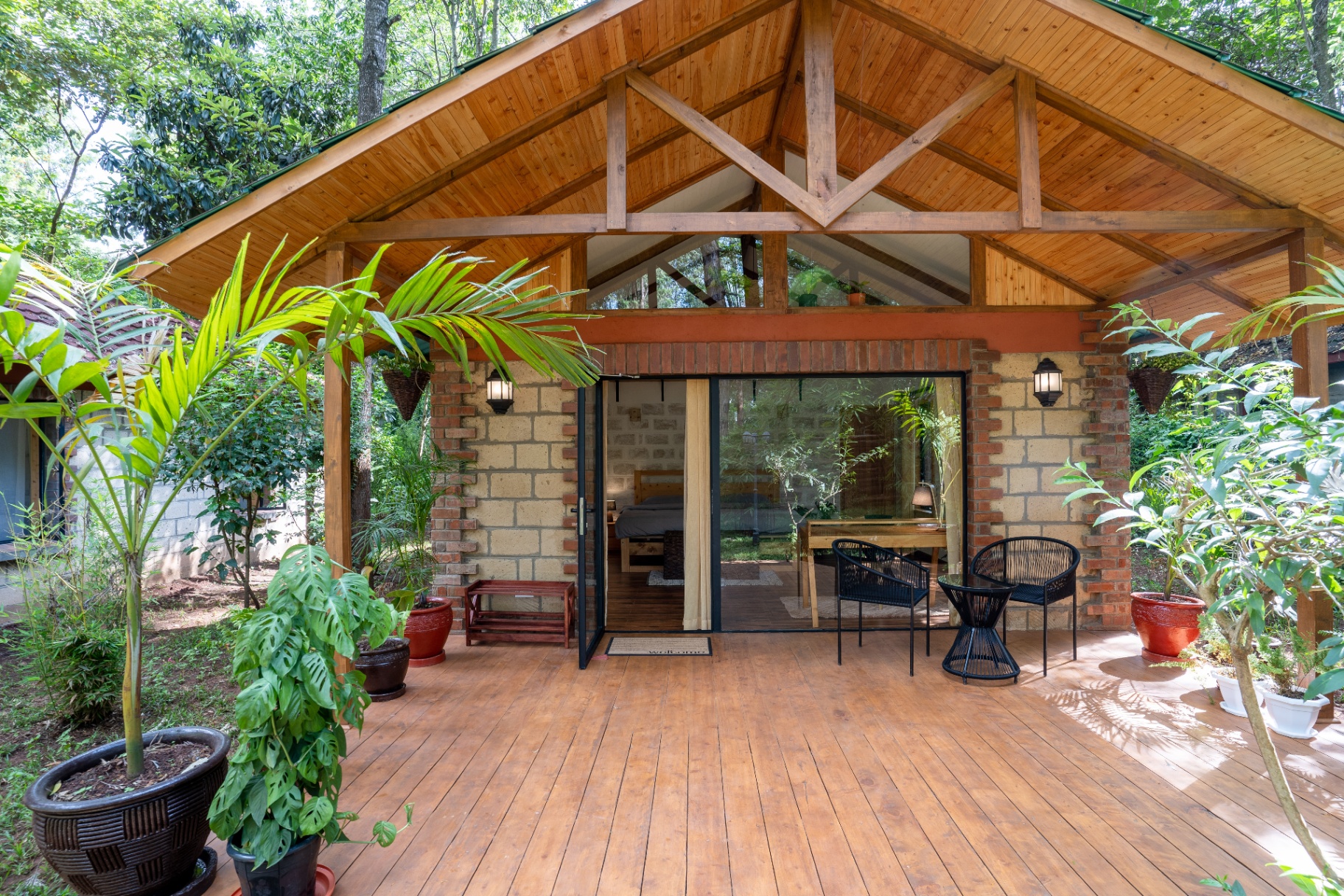 Front elevation of Cottage 1 with pitched cedar roof, exposed timber trusses, brick-and-stone walls and a wooden deck framed by palms and monstera