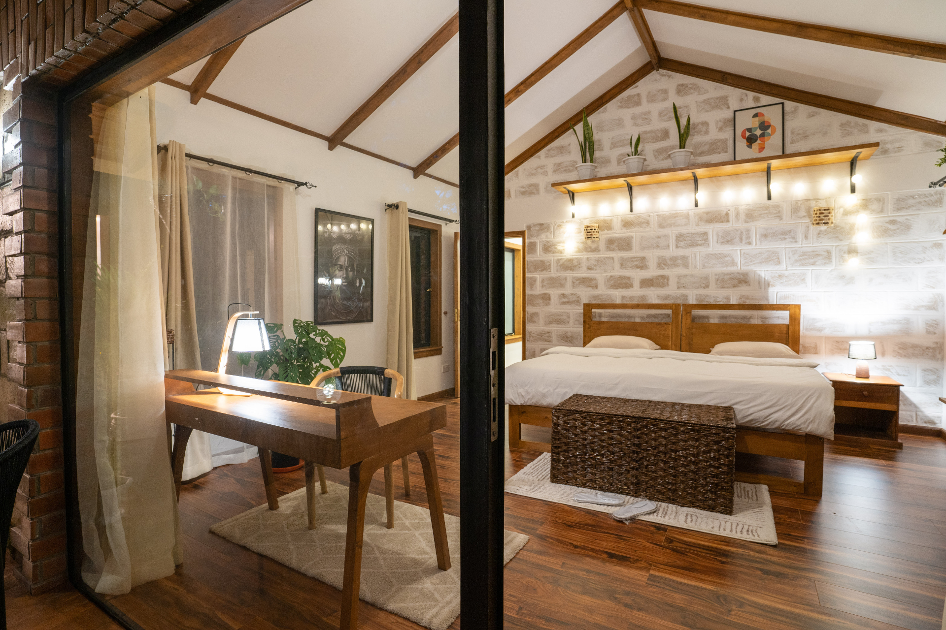 Nighttime view into Cottage 2 bedroom showing a timber writing desk by the glass doors, warm lamplight, wicker trunk and the bed against a softly lit stone wall