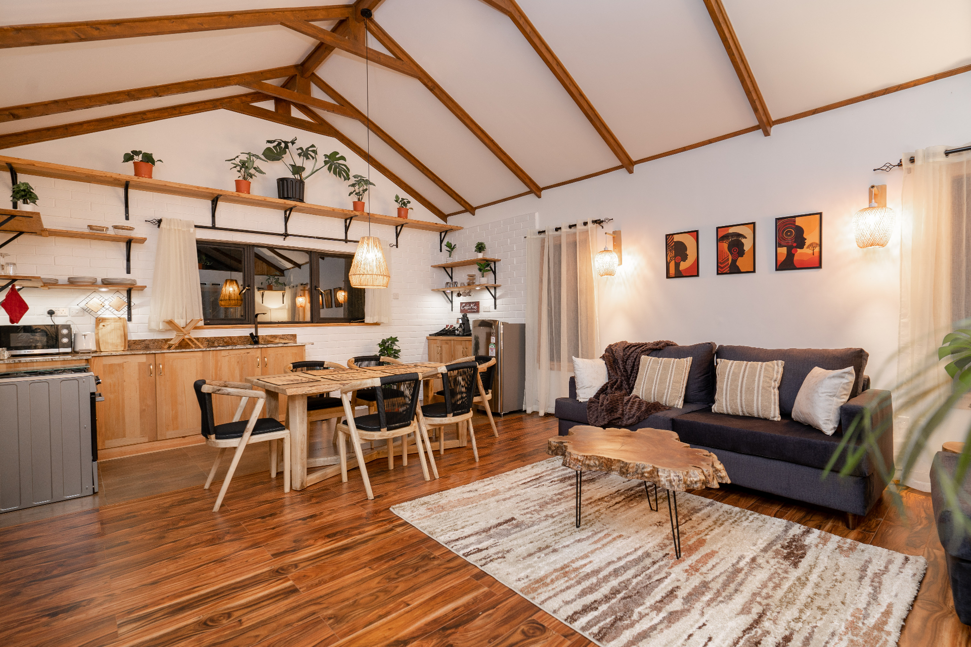 Wide evening view of Cottage 1 showing the open kitchen, dining table and sofa together under exposed timber beams and layered shelves with potted plants
