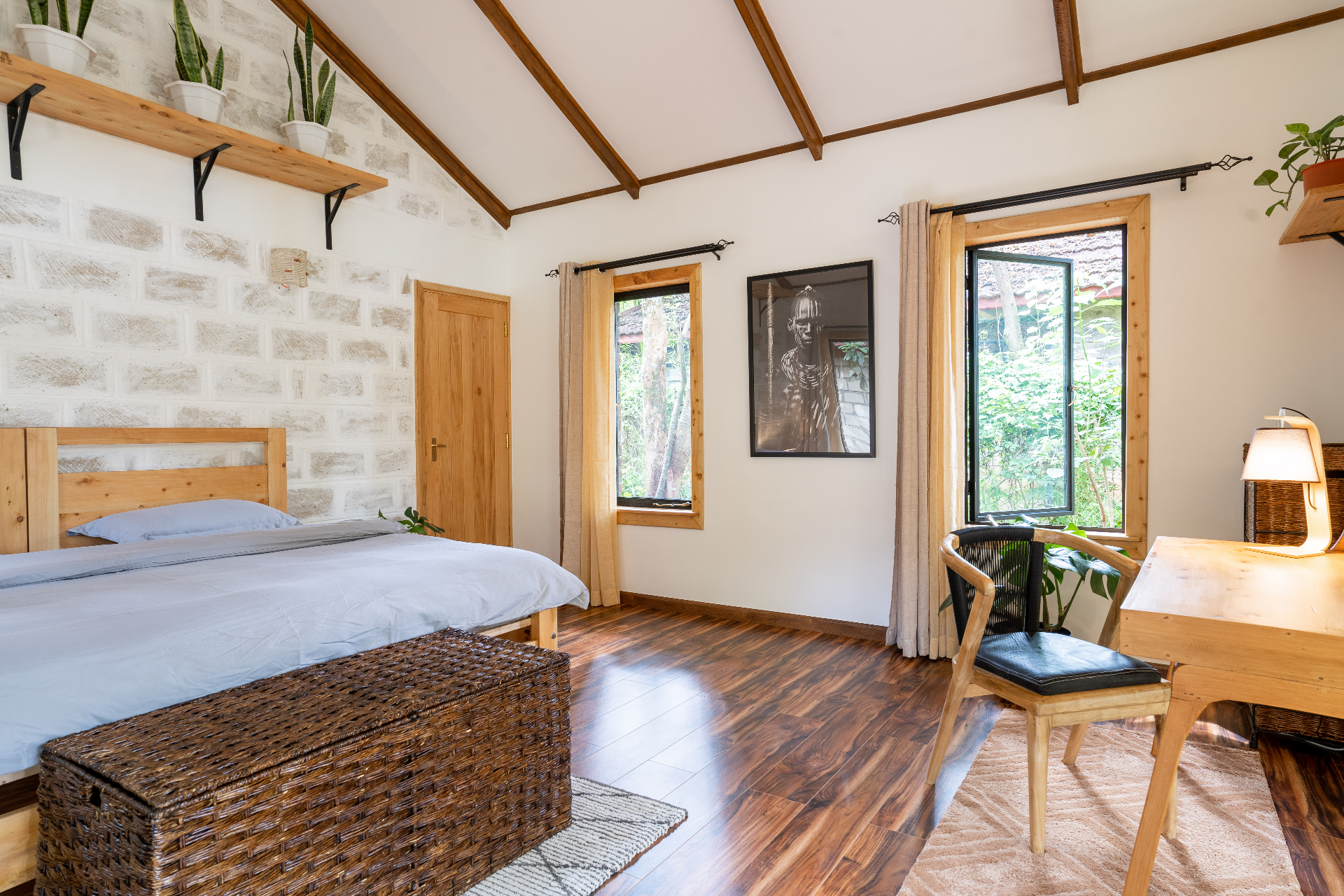 Bedroom view across the bed toward a timber writing desk, woven bench and two tall windows overlooking the garden