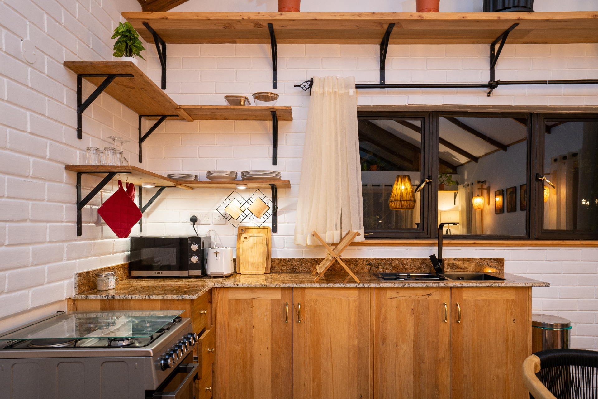Close-up of the Cottage 1 kitchen with granite worktop, gas cooker, microwave, toaster, chopping boards, open timber shelving and a black tap over the sink beside a window