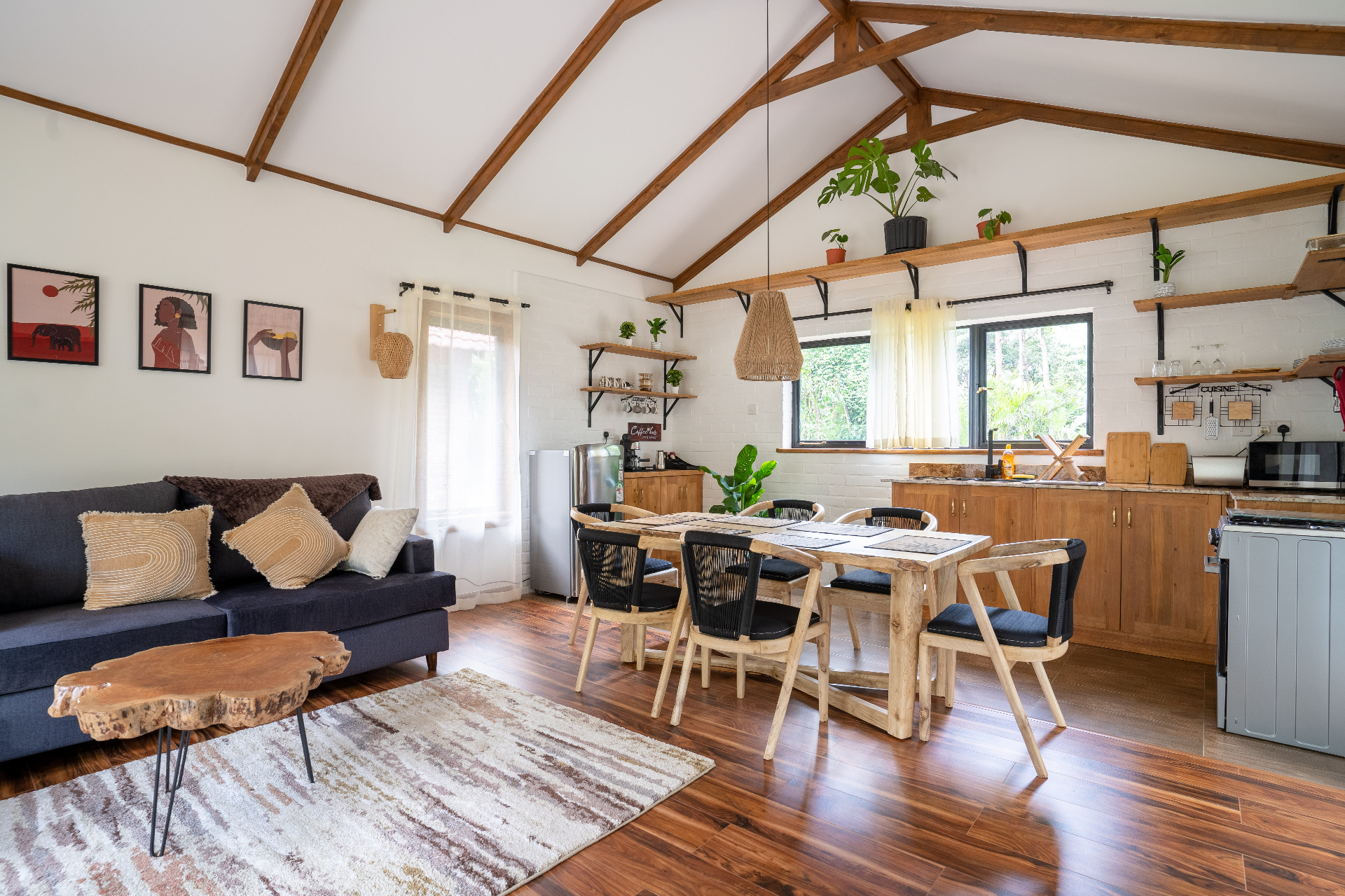 Wide daytime view across Cottage 2 showing the lounge sofa, live-edge coffee table, dining table, kitchen cabinetry and vaulted ceiling with exposed timber trusses