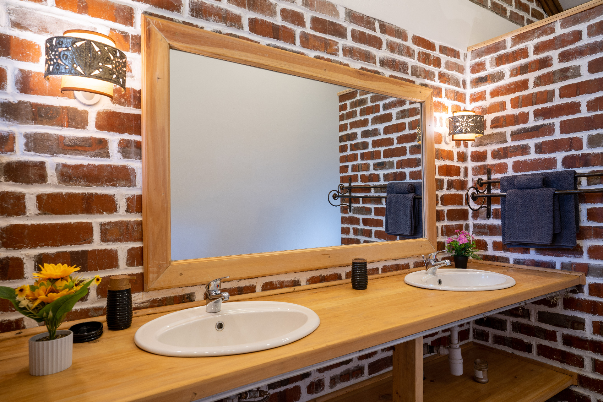Twin white basins set into a long timber counter under a large framed mirror, flanked by patterned metal sconces on a whitewashed brick wall