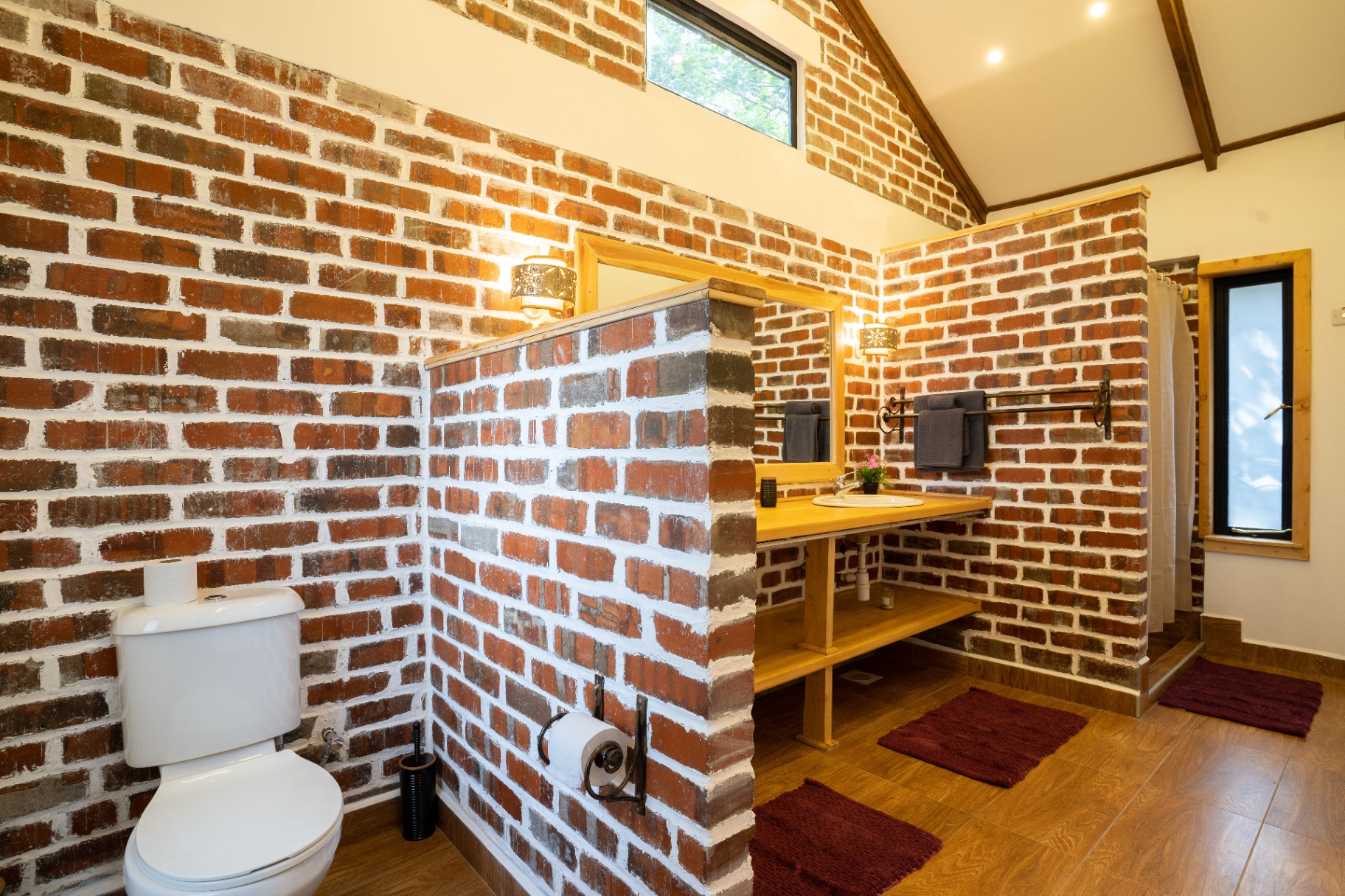 Wide view of the exposed-brick bathroom with a half-height brick divider between the toilet and the timber vanity, walk-in shower and burgundy bath mats on timber floors