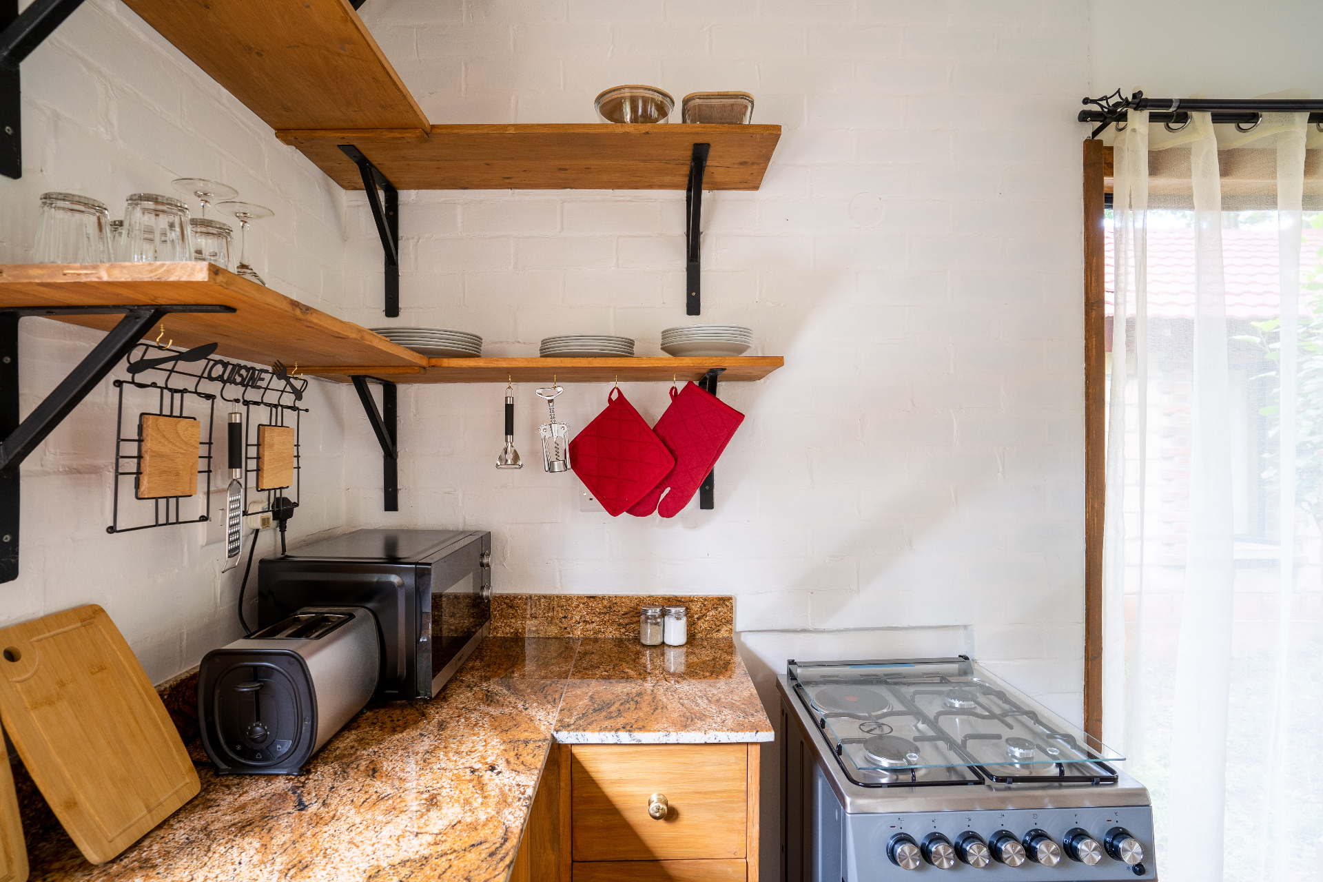 Close-up of Cottage 2 kitchen showing granite countertop, gas stove, red oven mitts, hanging utensils and stacked white plates on open timber shelves against a whitewashed brick wall