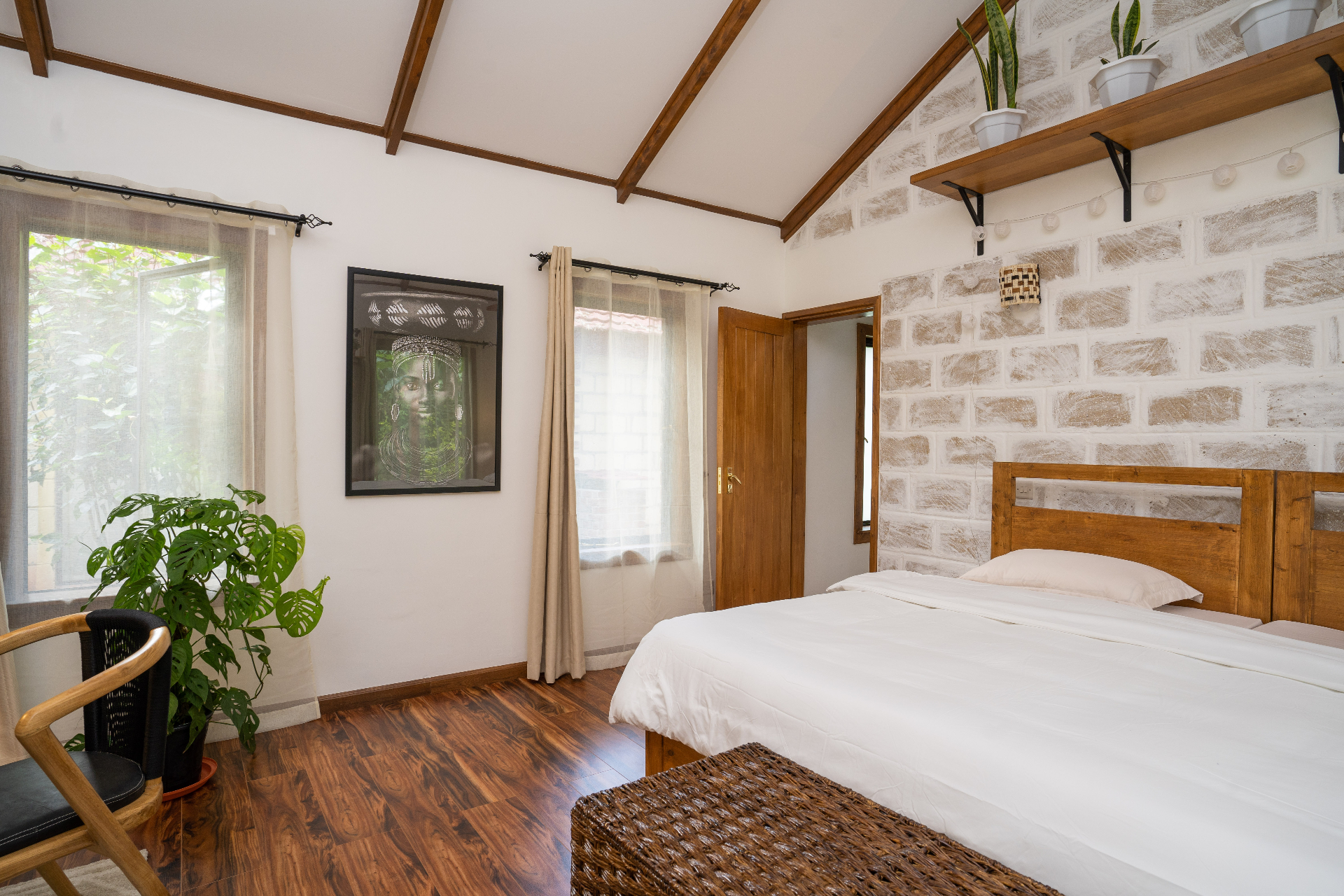 Side angle of Cottage 2 bedroom showing the timber bed, hardwood floor, sheer curtains at the windows, framed African portrait and layered natural textures