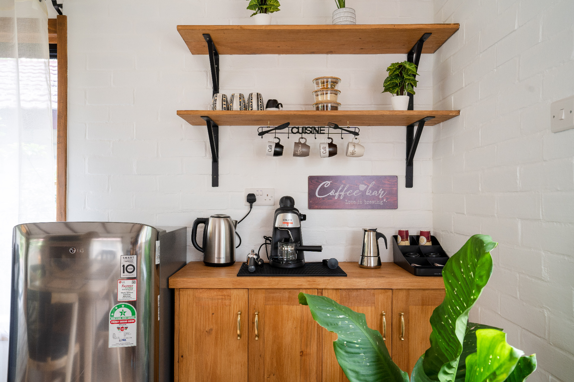 Cottage 2 coffee bar with kettle, espresso maker, moka pot and capsule tray on a timber cabinet, under open shelves of mugs and jars with a Coffee Bar sign on the brick wall