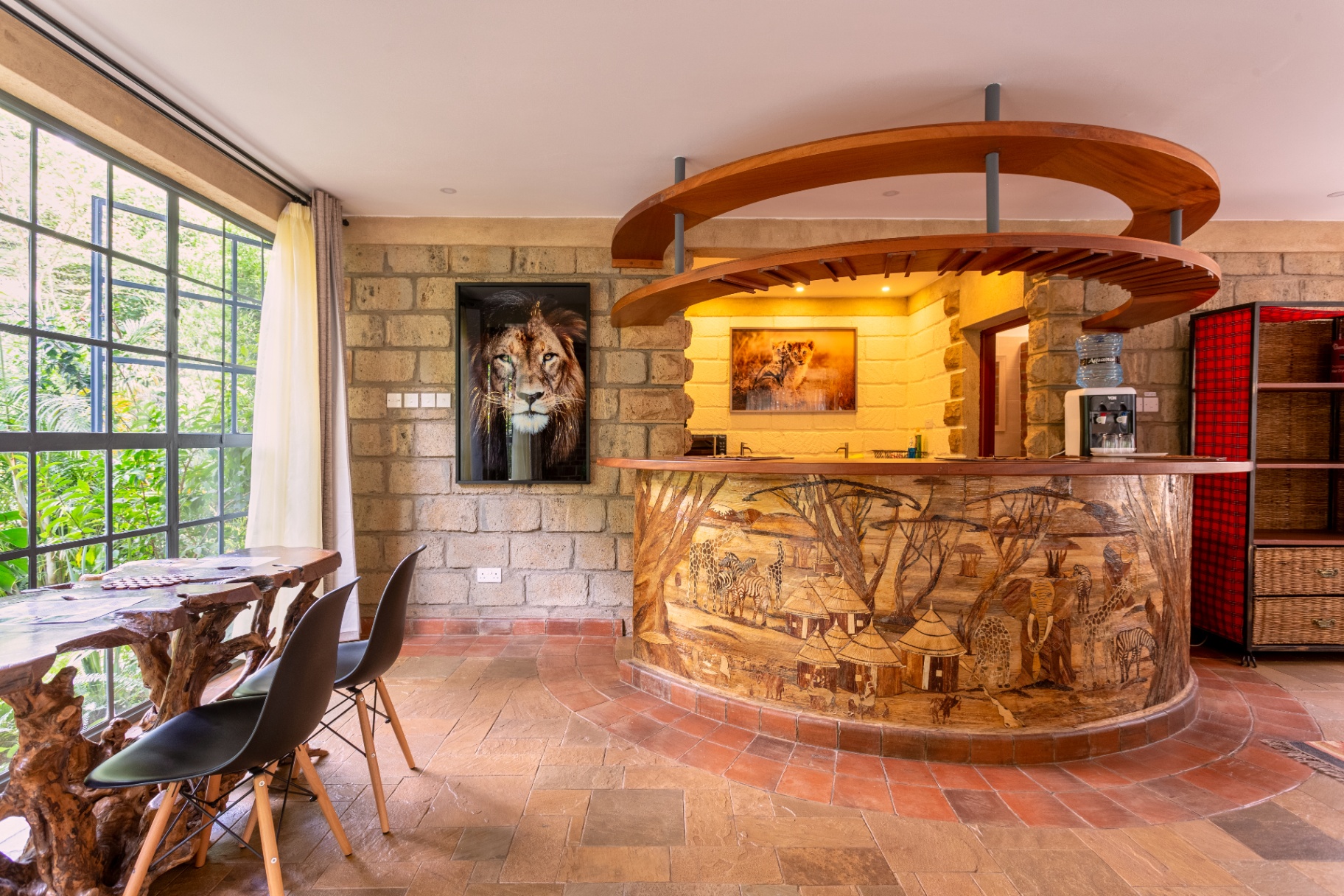 Wide view of the curved carved-wood safari bar and lion portrait with the root-carved dining table beside the garden window